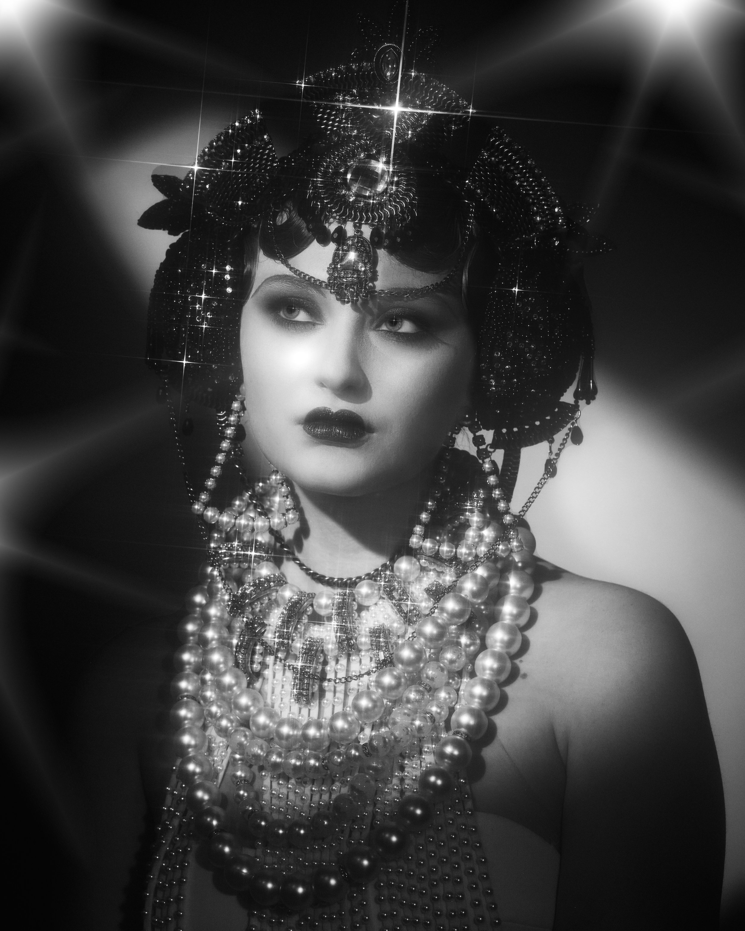A black-and-white portrait of a woman wearing elaborate jewelry, a headpiece, and dark lipstick, with a serious expression.