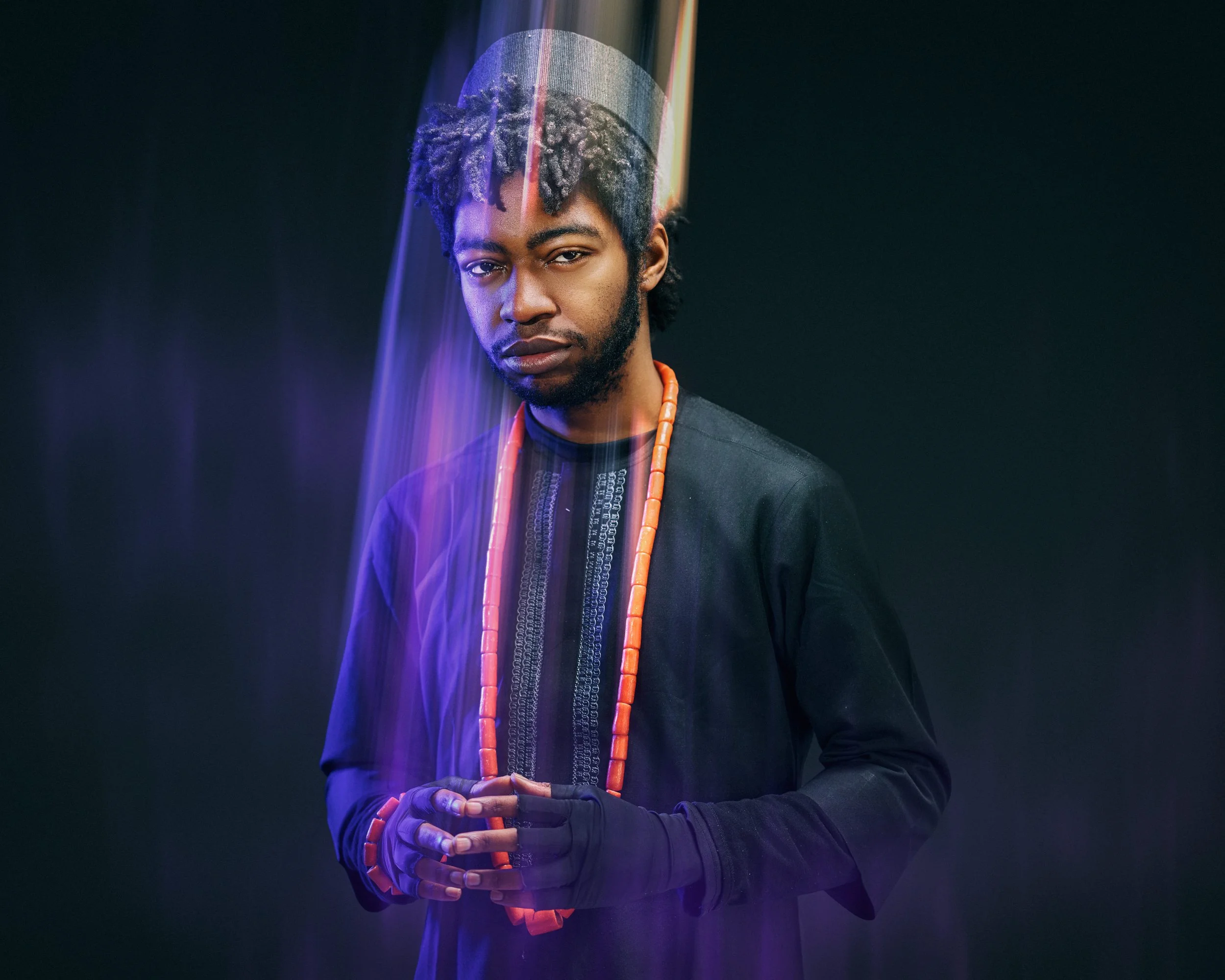 A young man with braided hair and a beard, wearing a black traditional African shirt and orange beaded necklace, stands against a dark background with colorful light streaks.