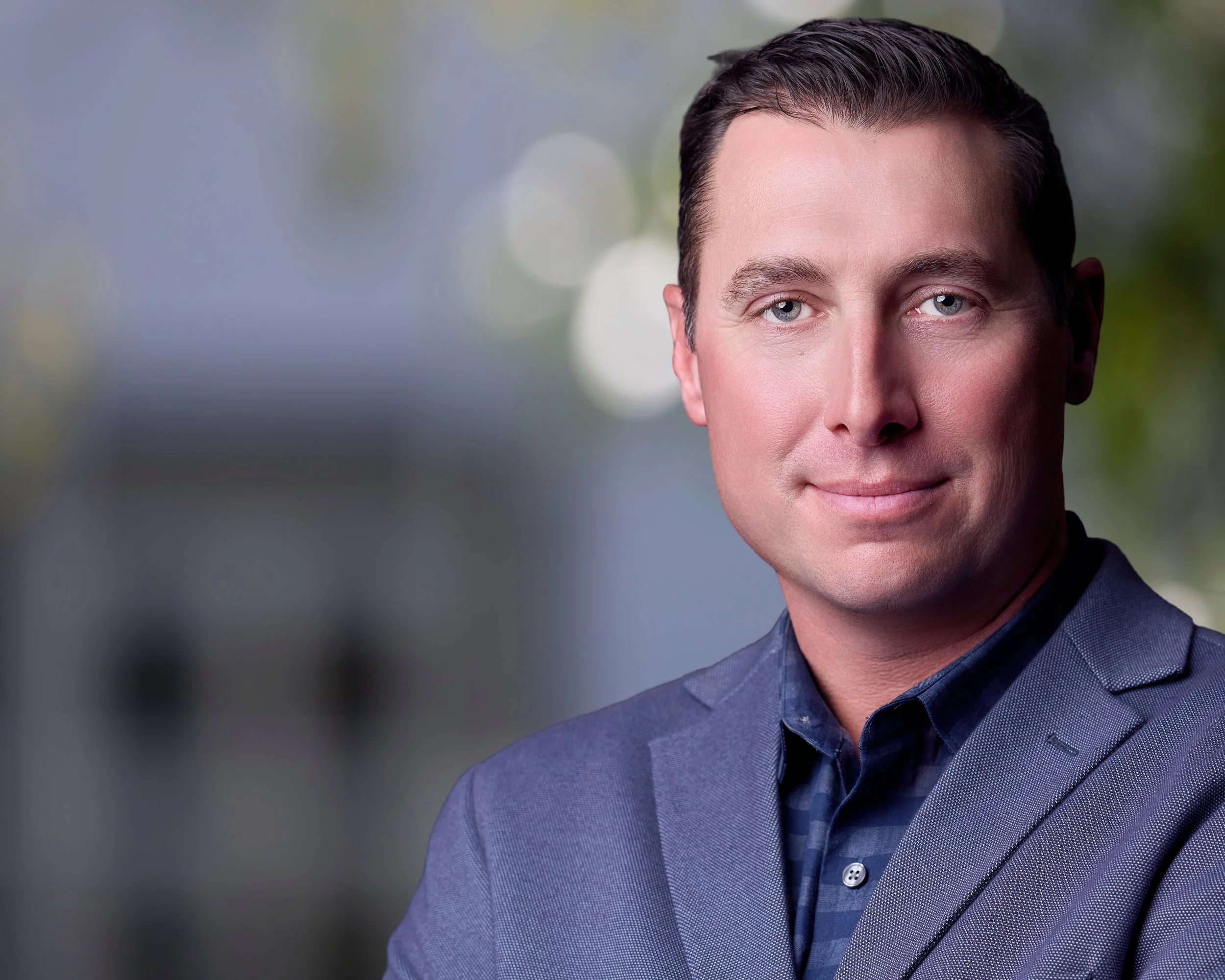 A man with short dark hair, blue eyes, and a slight smile, wearing a blue suit jacket and a dark blue shirt, standing outdoors with a blurred background of greenery.