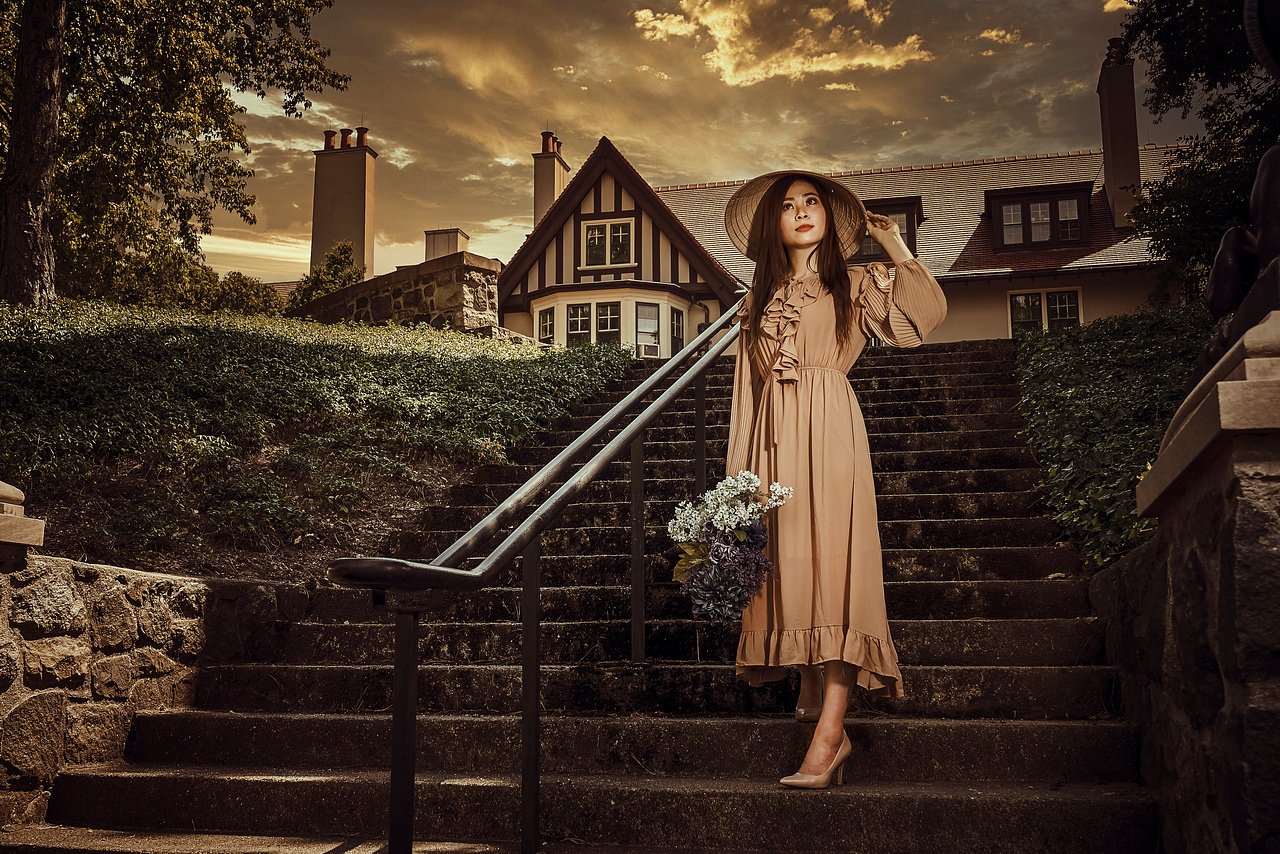 A woman in vintage style dress holding a bouquet and hat, standing on outdoor stairs in front of a large house with chimney, under a dramatic sunset sky.
