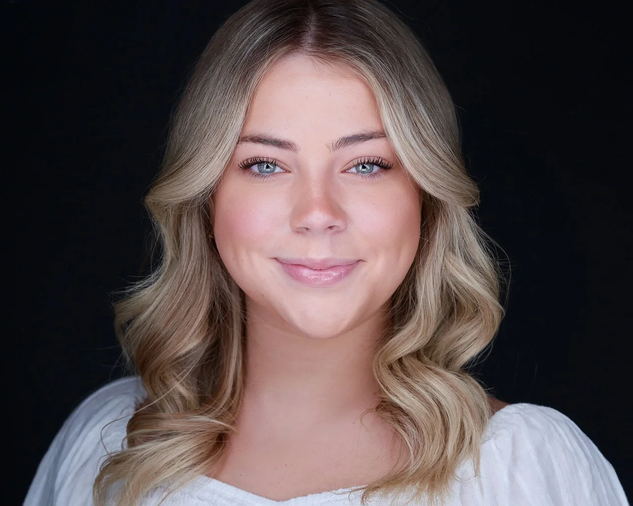 Close-up portrait of a young woman with blonde hair, blue eyes, and light makeup, smiling slightly against a black background.