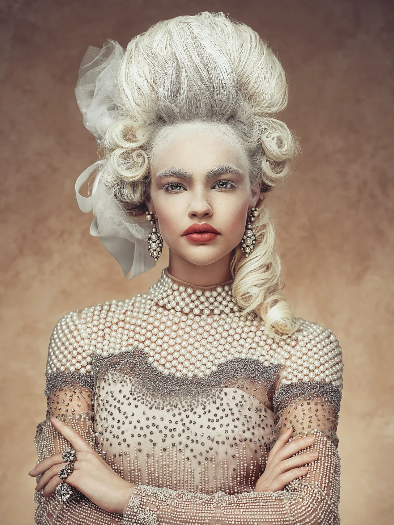 A woman with platinum blonde hair styled in an elaborate vintage updo, wearing pearl earrings, a pearl-embellished dress, and ornate rings, posed with arms crossed against a neutral background.