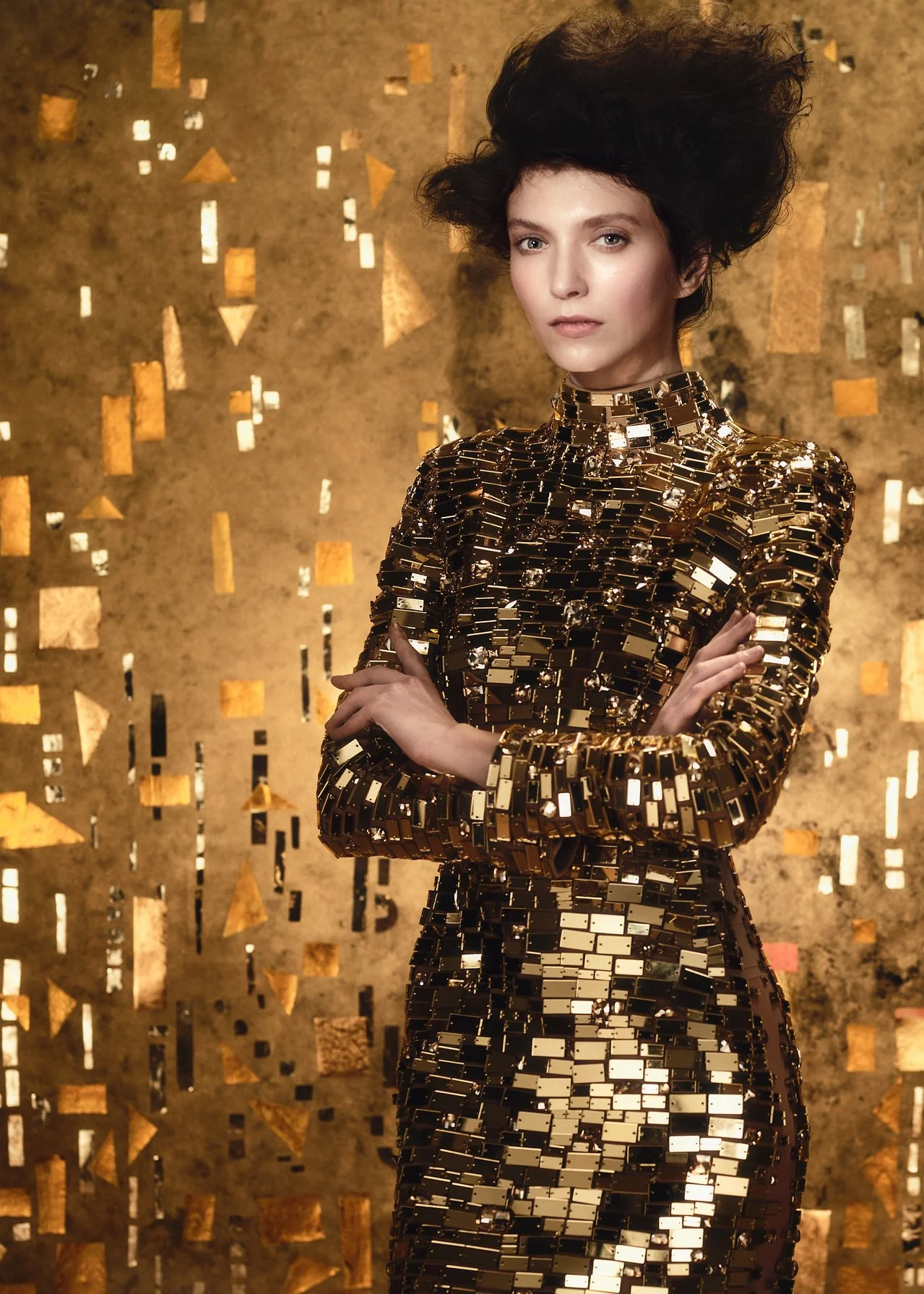A woman in a form-fitting dress made of shiny, rectangular, reflective gold tiles stands with her arms crossed against a gold background with scattered geometric shapes.