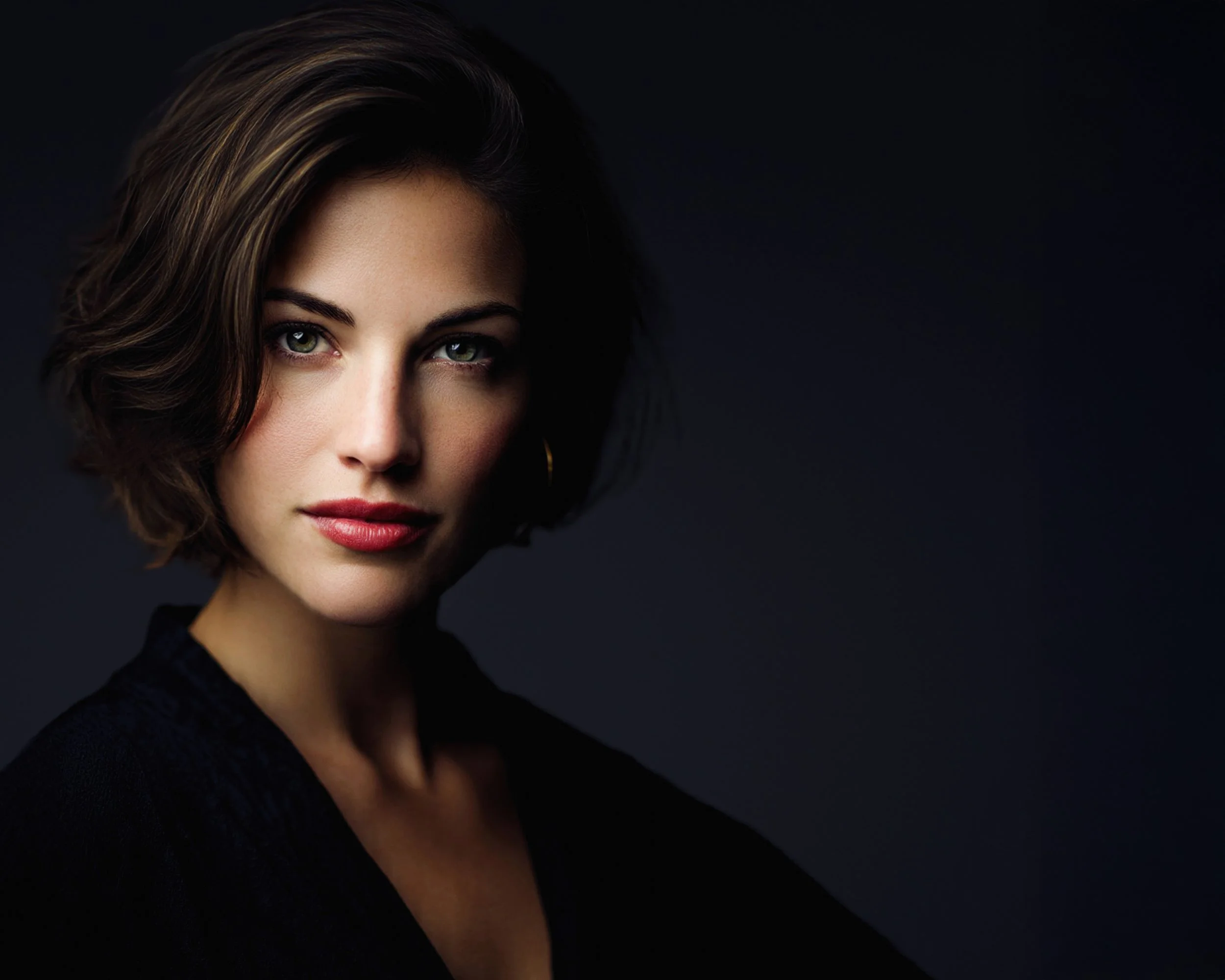 A woman with short wavy brown hair and green eyes, wearing a black top and gold hoop earrings, looking directly at the camera against a dark background.