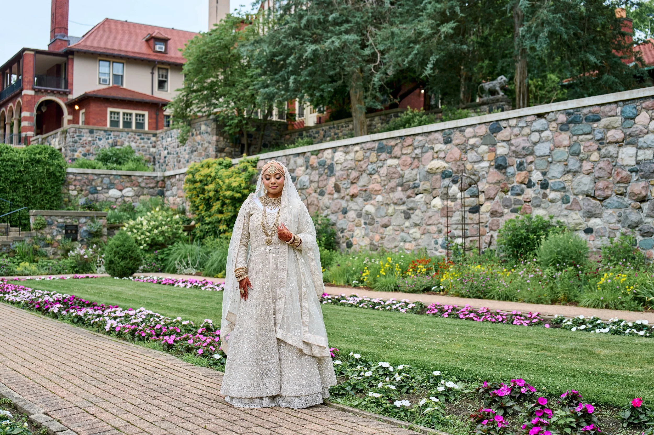A woman dressed in a cream-colored traditional wedding outfit with jewelry, standing on a brick pathway in a garden with flowers and greenery, with a stone wall and house in the background.
