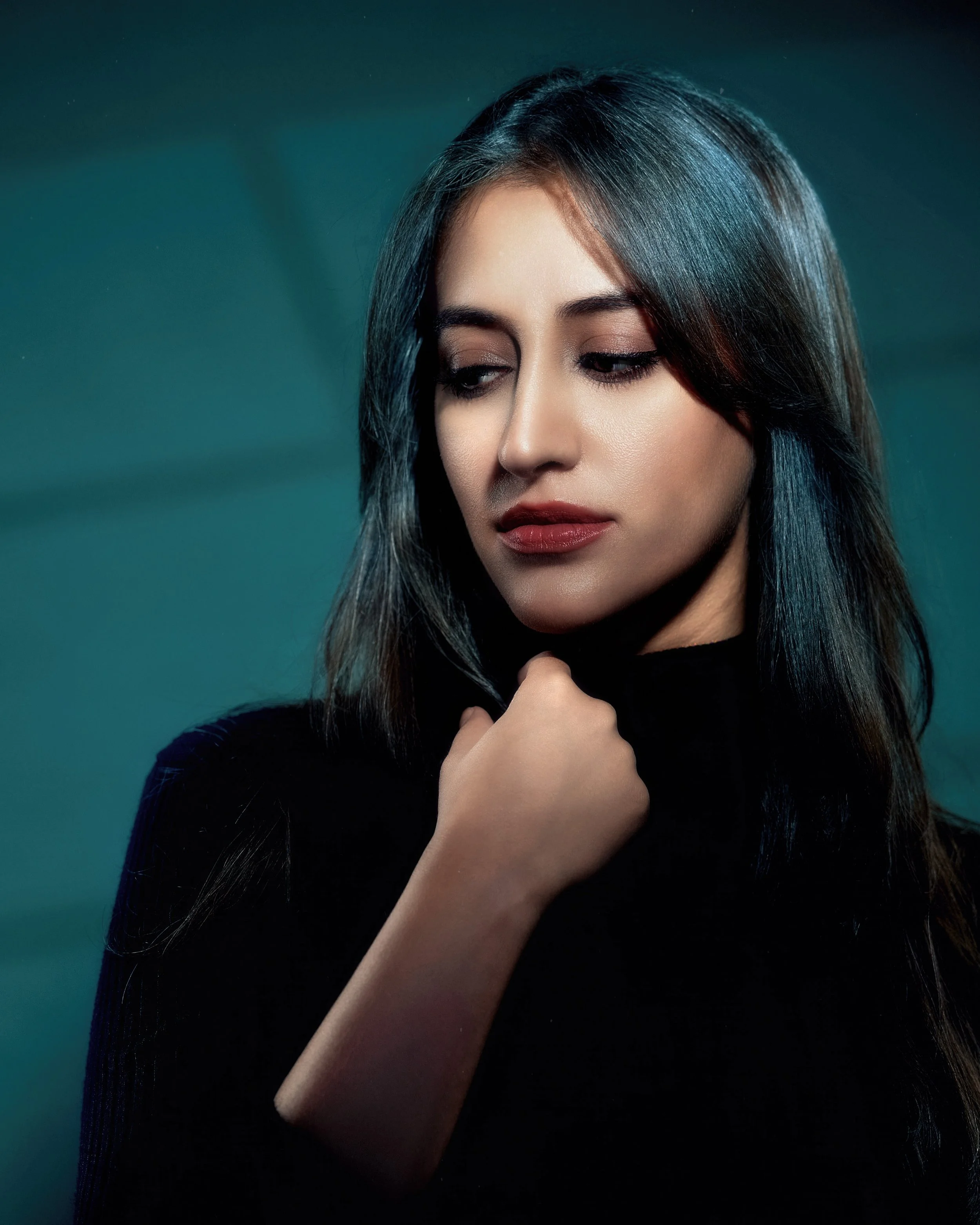 A woman with long dark hair, wearing a black top, looking down and slightly to the side with a thoughtful expression, against a dark teal background.