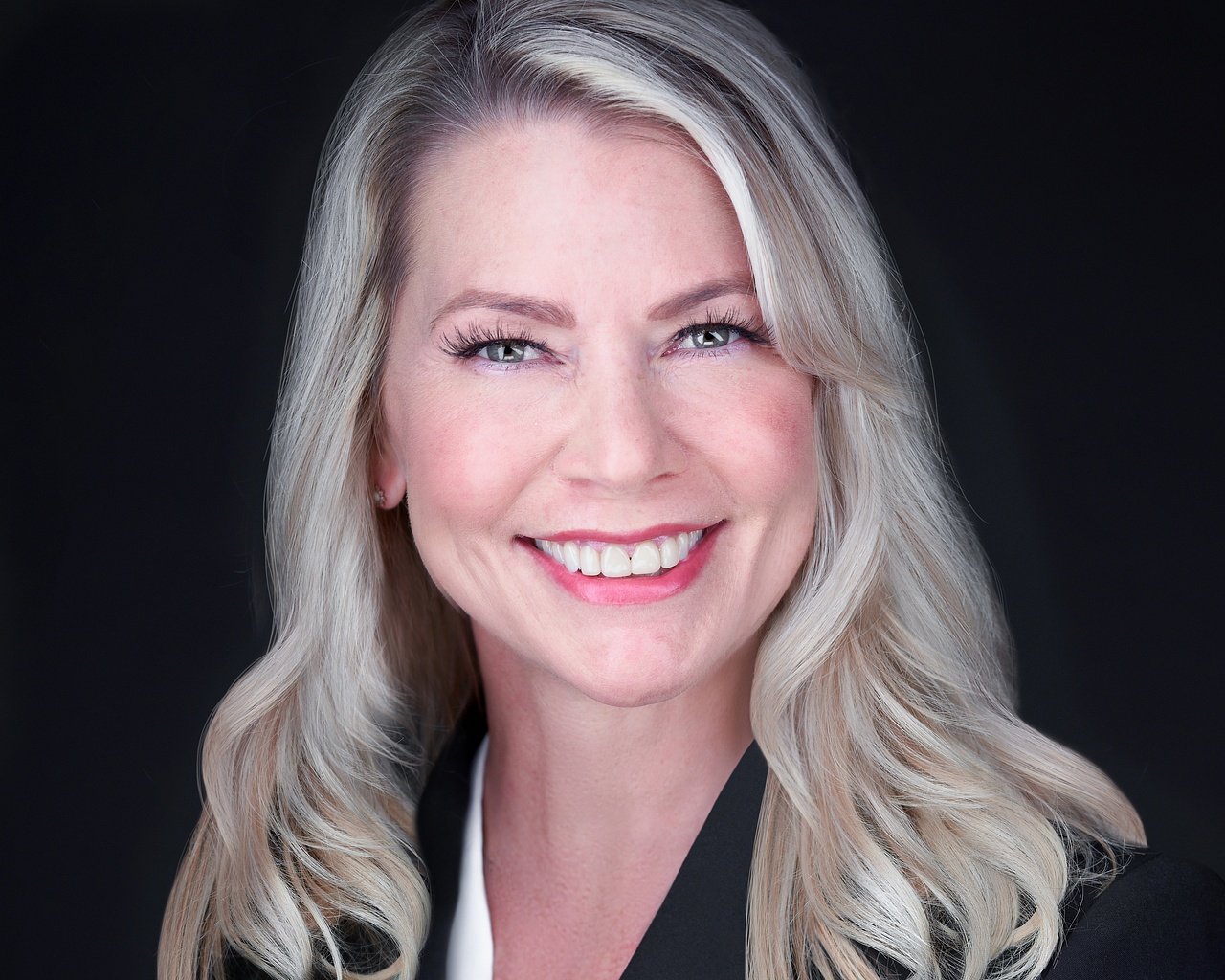 Headshot of a smiling blonde woman with light skin and blue eyes against a dark background.