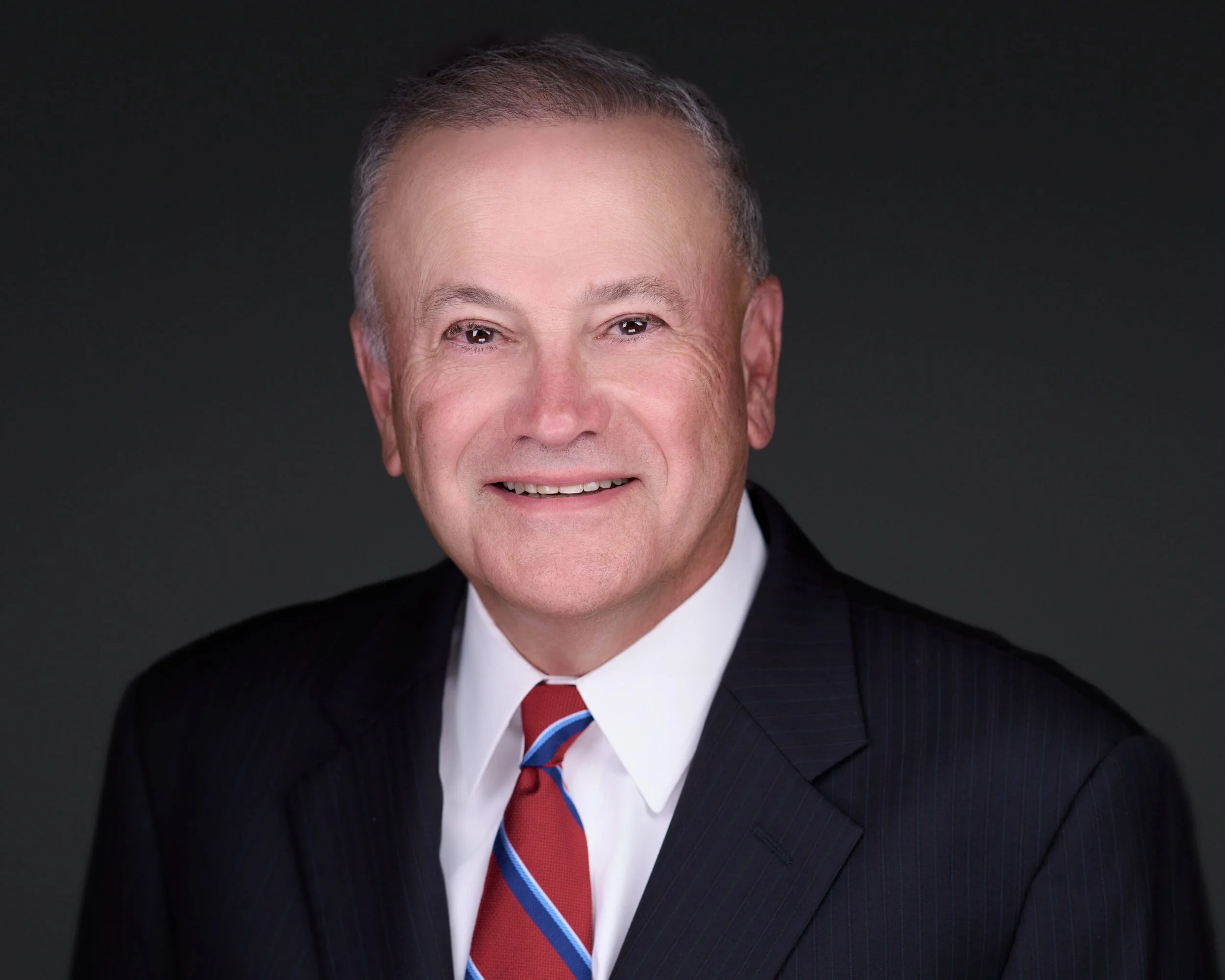 Portrait of a smiling middle-aged man wearing a business suit, white shirt, and a striped red and blue tie, against a dark background.
