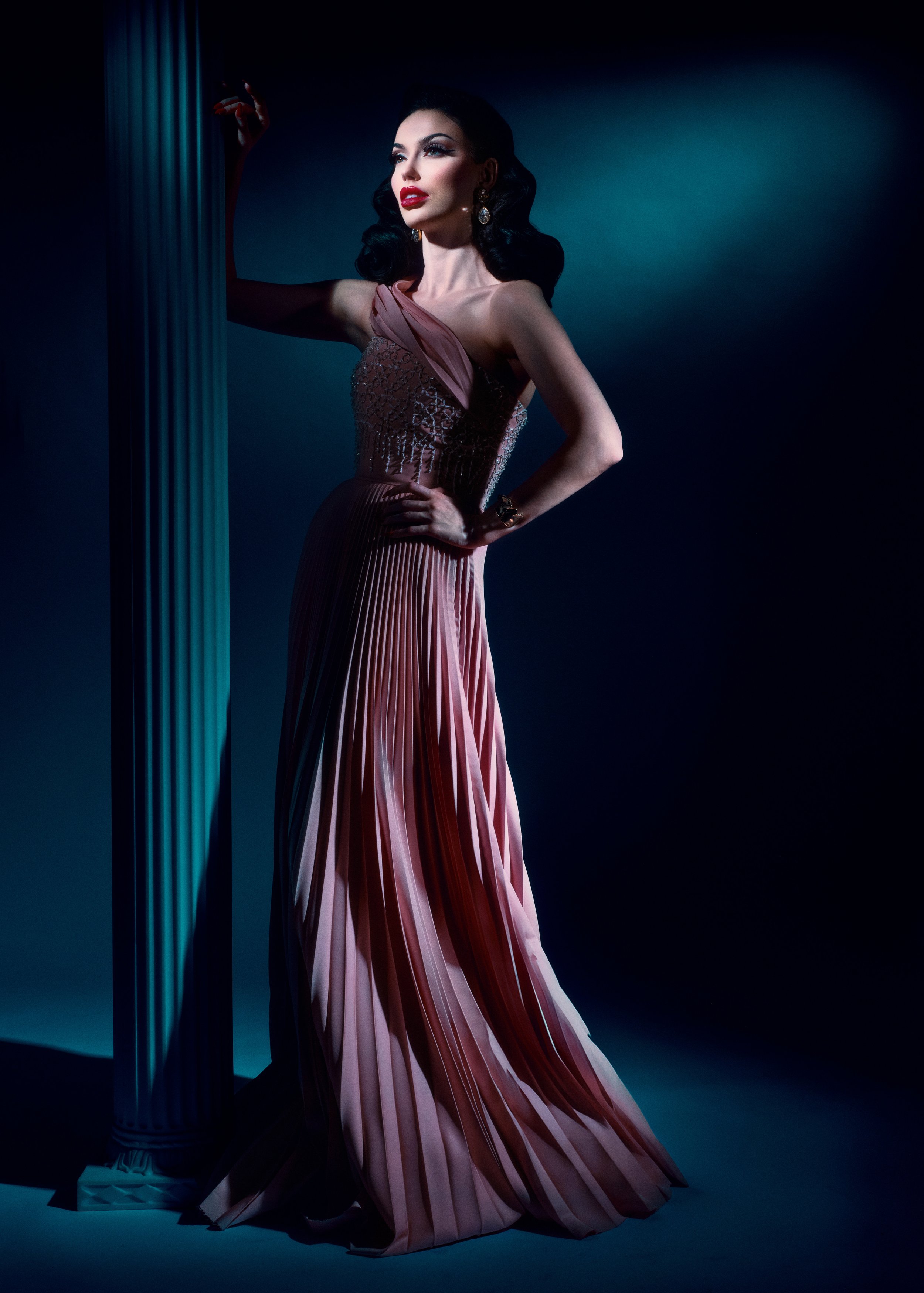 A woman in a glamorous, pink strapless evening gown with pleats stands beside a white classical column against a dark background.