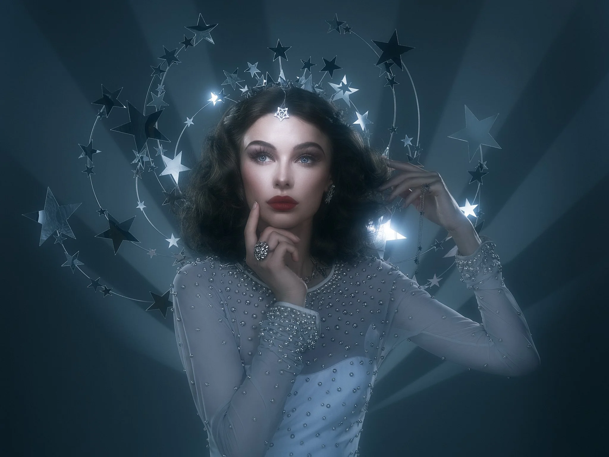 A woman with dark wavy hair and makeup, wearing a bejeweled sheer dress, posing with her hand on her chin and a hand holding jewelry. She is surrounded by a halo of silver stars against a dark background with light beams.