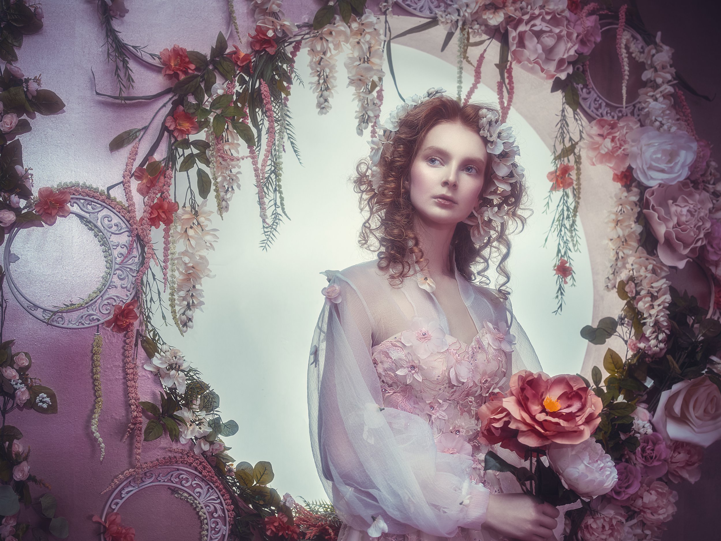 A woman with curly hair in a floral dress and a flower crown peers through a heart-shaped floral frame decorated with pink and purple flowers and greenery, holding a bouquet of large flowers.