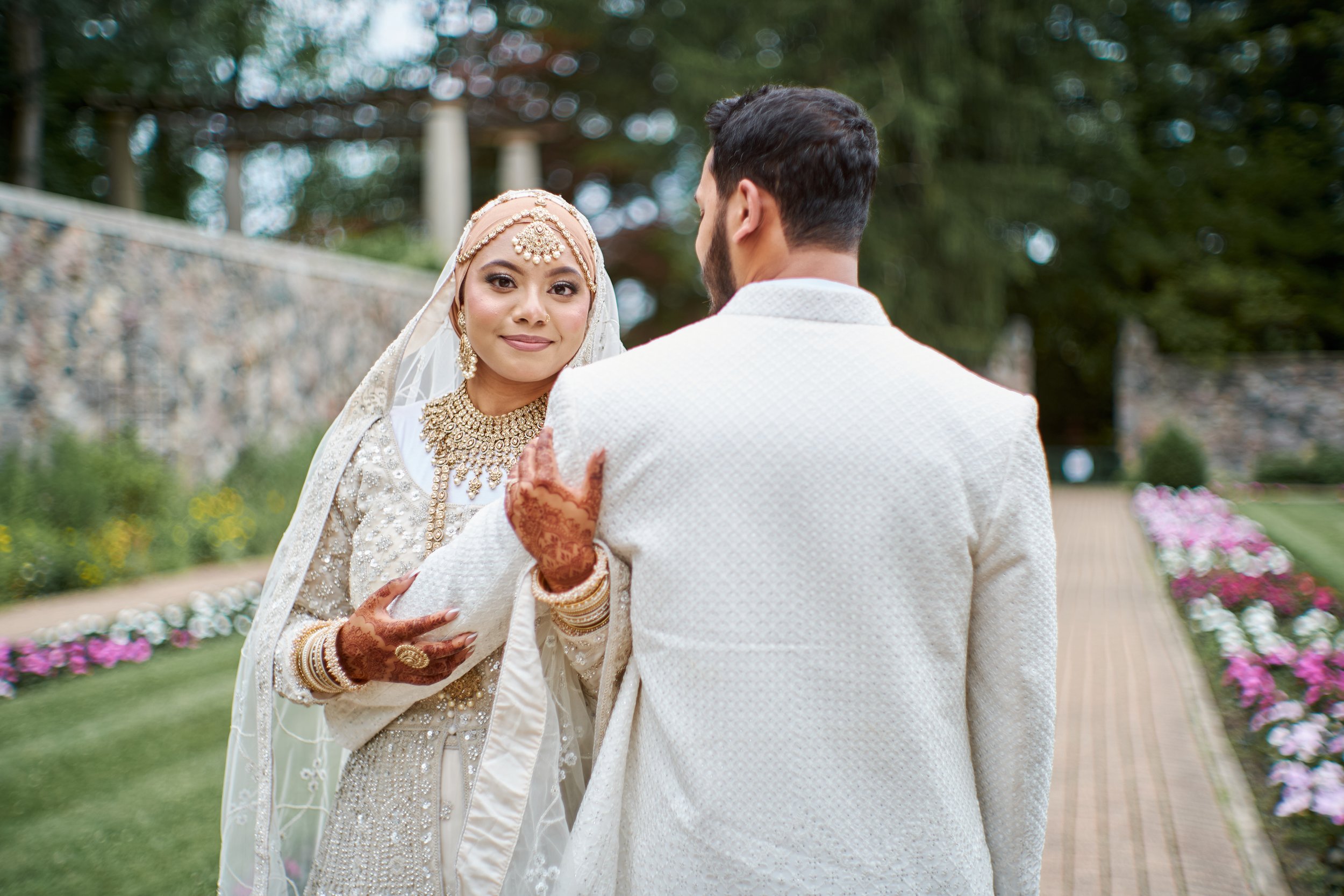 A bride in traditional wedding attire and a groom in a white suit standing outdoors on a garden pathway with flowers and greenery.