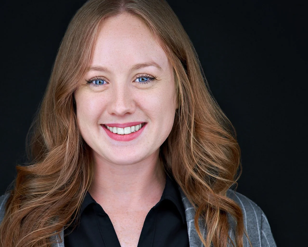 Close-up portrait of a smiling woman with long curly red hair, blue eyes, and fair skin, wearing a dark shirt and gray blazer, against a black background.