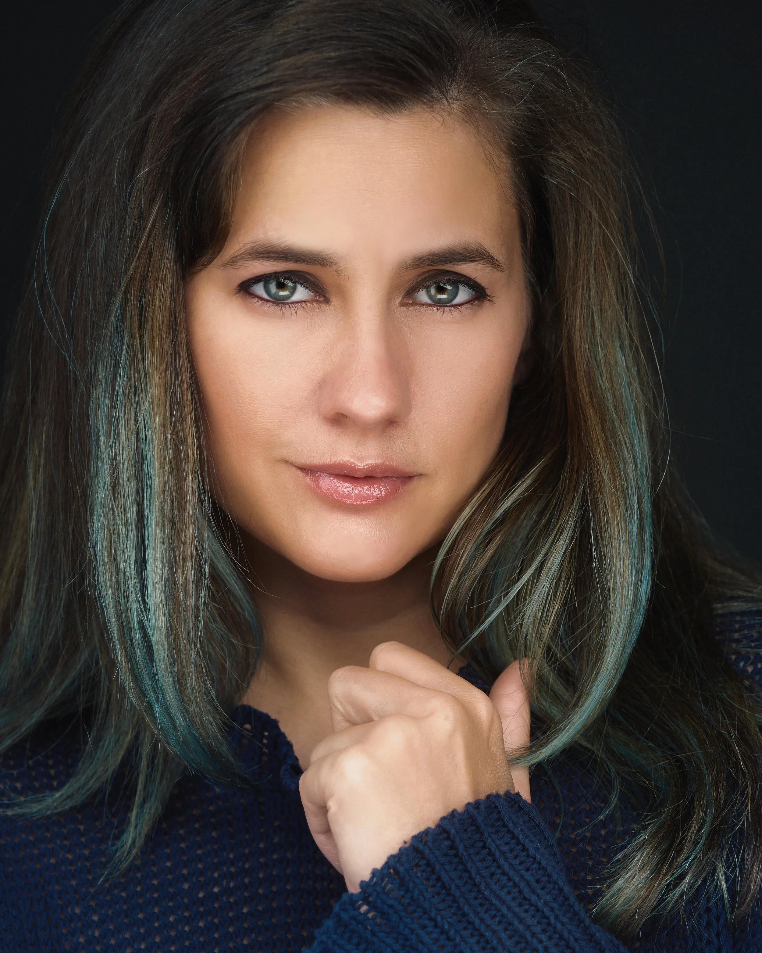 Close-up portrait of a woman with blue eyes and long brown hair with green highlights, looking directly at the camera. She has a neutral expression and is wearing a dark blue sweater, with her hand gently touching her chin.