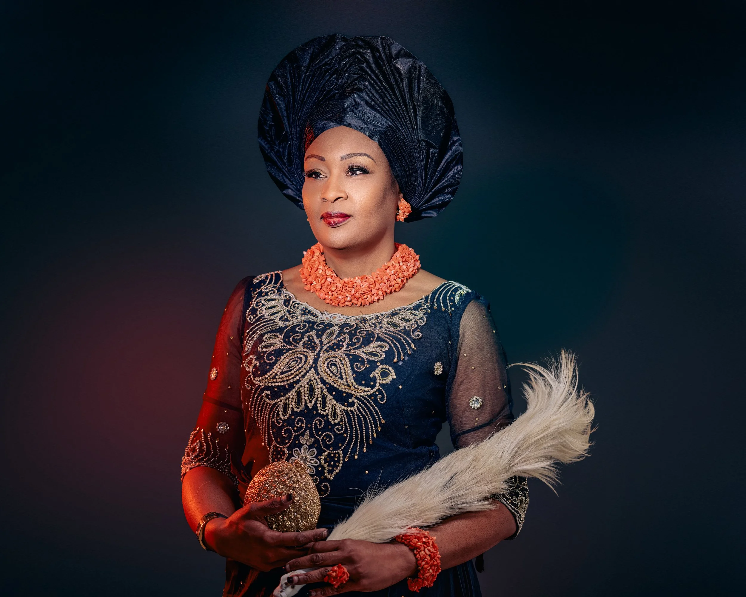 A woman dressed in traditional African attire, wearing a navy blue gele headwrap, ornate embroidered dress, and coral jewelry, holding a feather and a beaded clutch against a dark background.