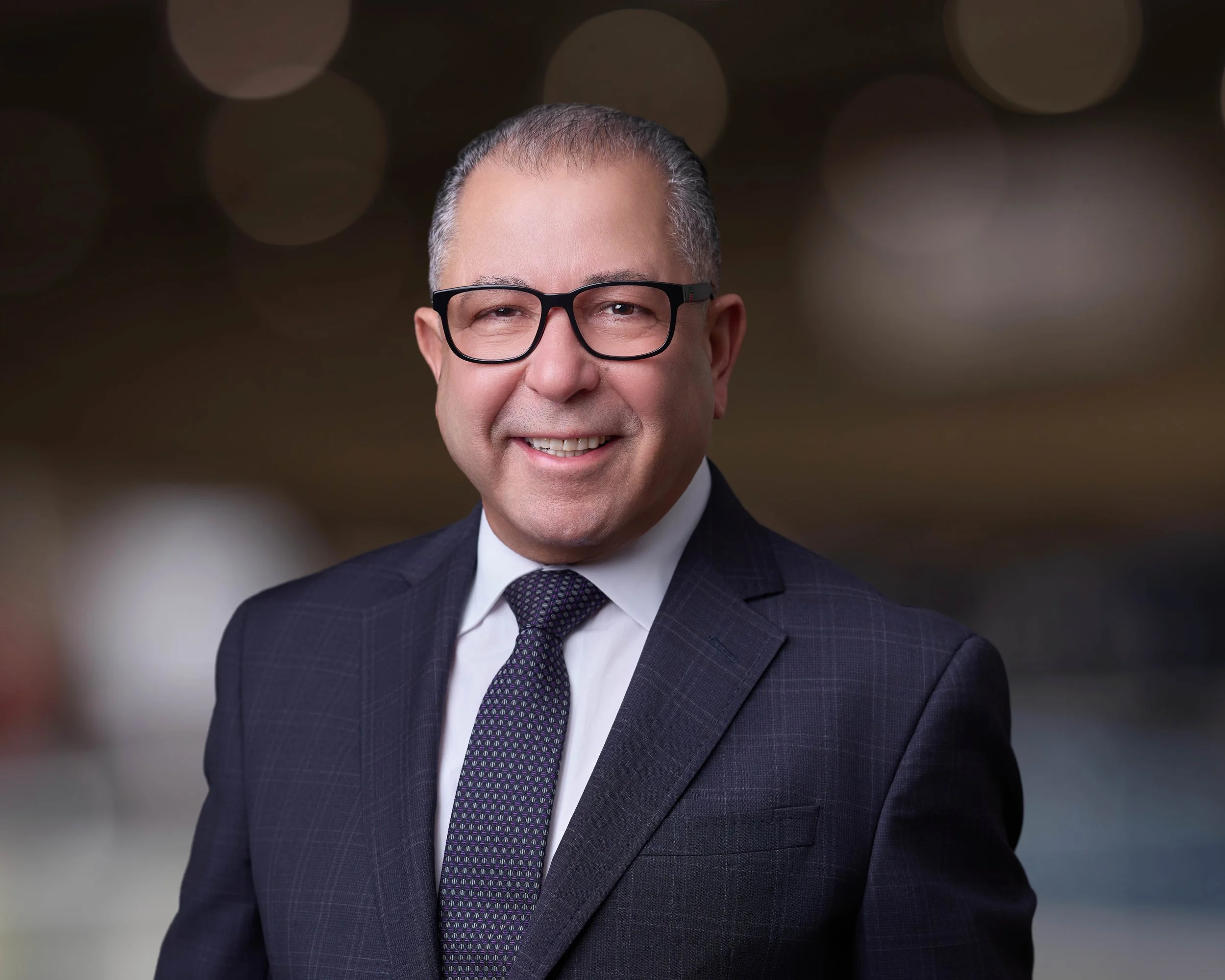 A middle-aged man with glasses prays in a business suit with a white shirt and patterned tie, smiling against a blurred background with circular light bokeh.