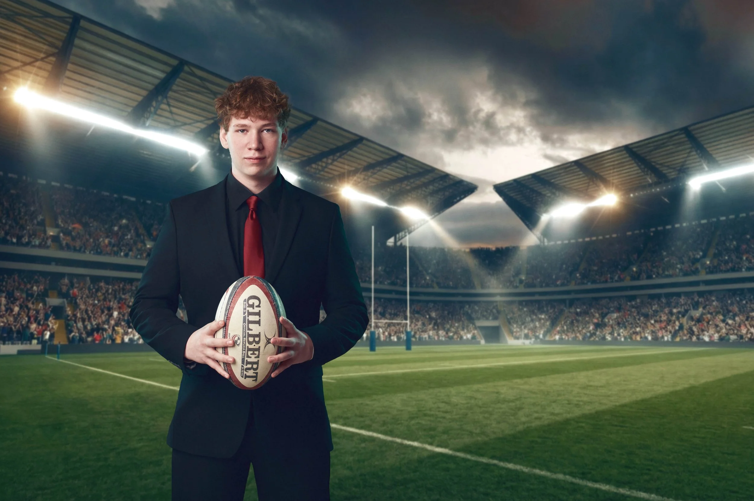 A young man in a black suit and red tie holding a rugby ball stands in a rugby stadium filled with spectators under a cloudy sky.