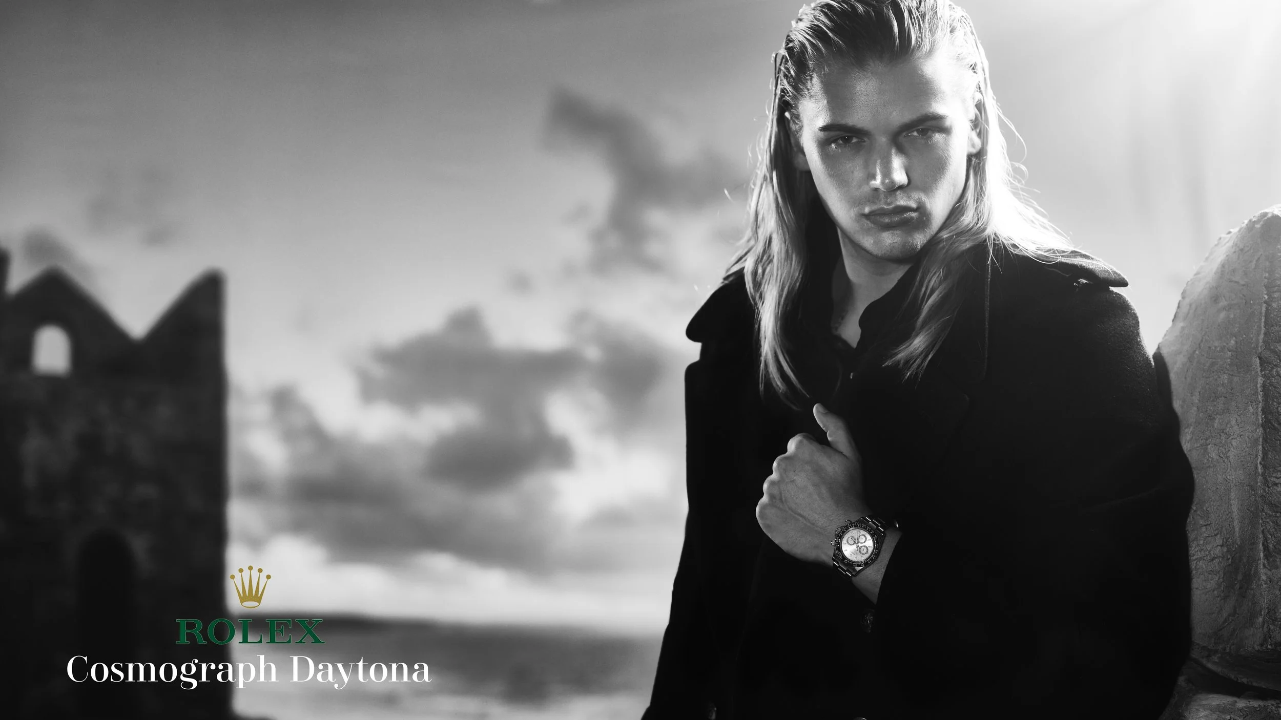 Black and white photo of a young man with long hair wearing a watch and a dark jacket, standing outdoors near a stone structure, with a cloudy sky in the background. Text overlay includes the Rolex logo and the words "Cosmograph Daytona."