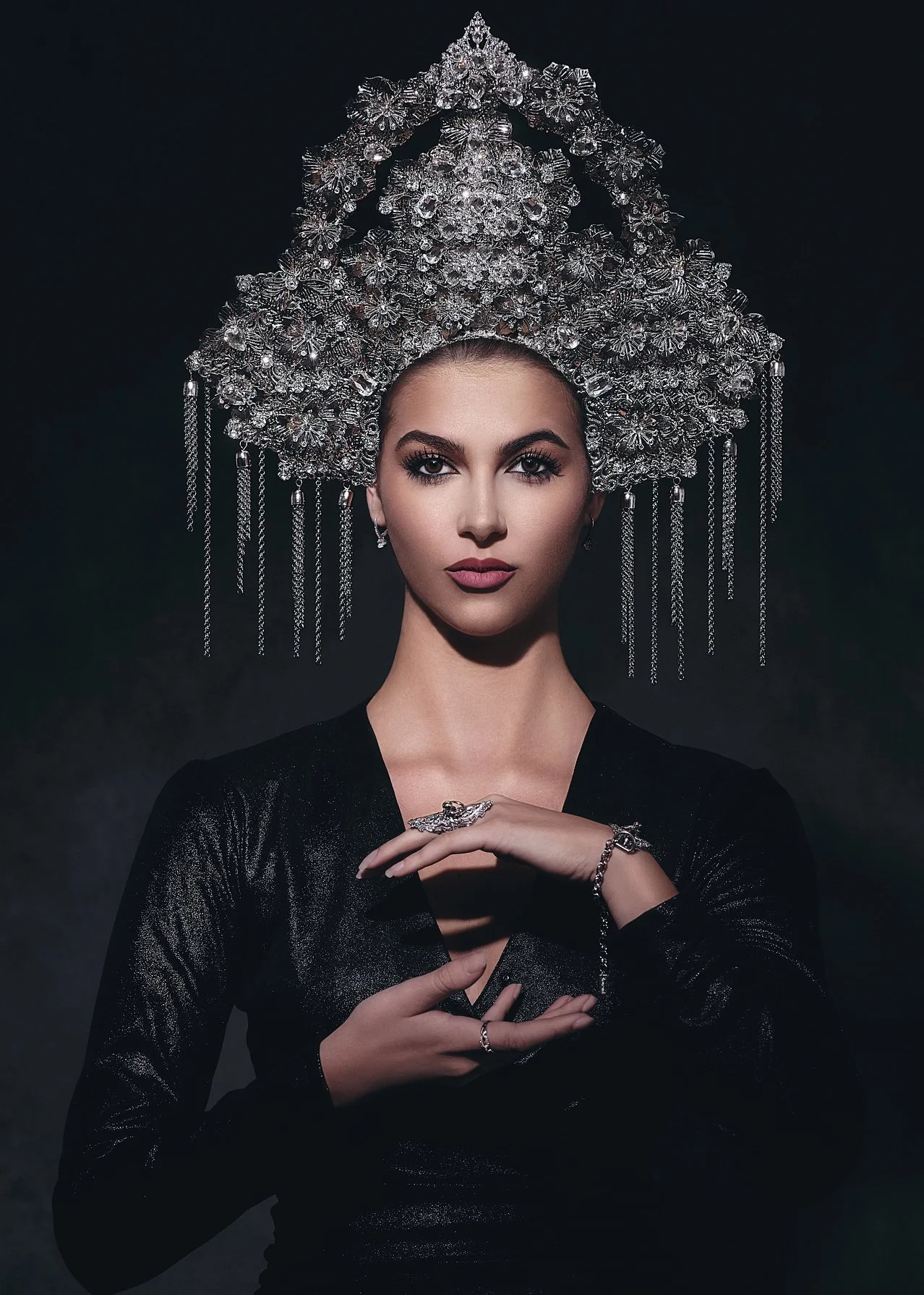 Portrait of a woman with a large ornate silver headpiece, wearing black shimmery clothing, jewelry, and looking directly at the camera against a dark background.