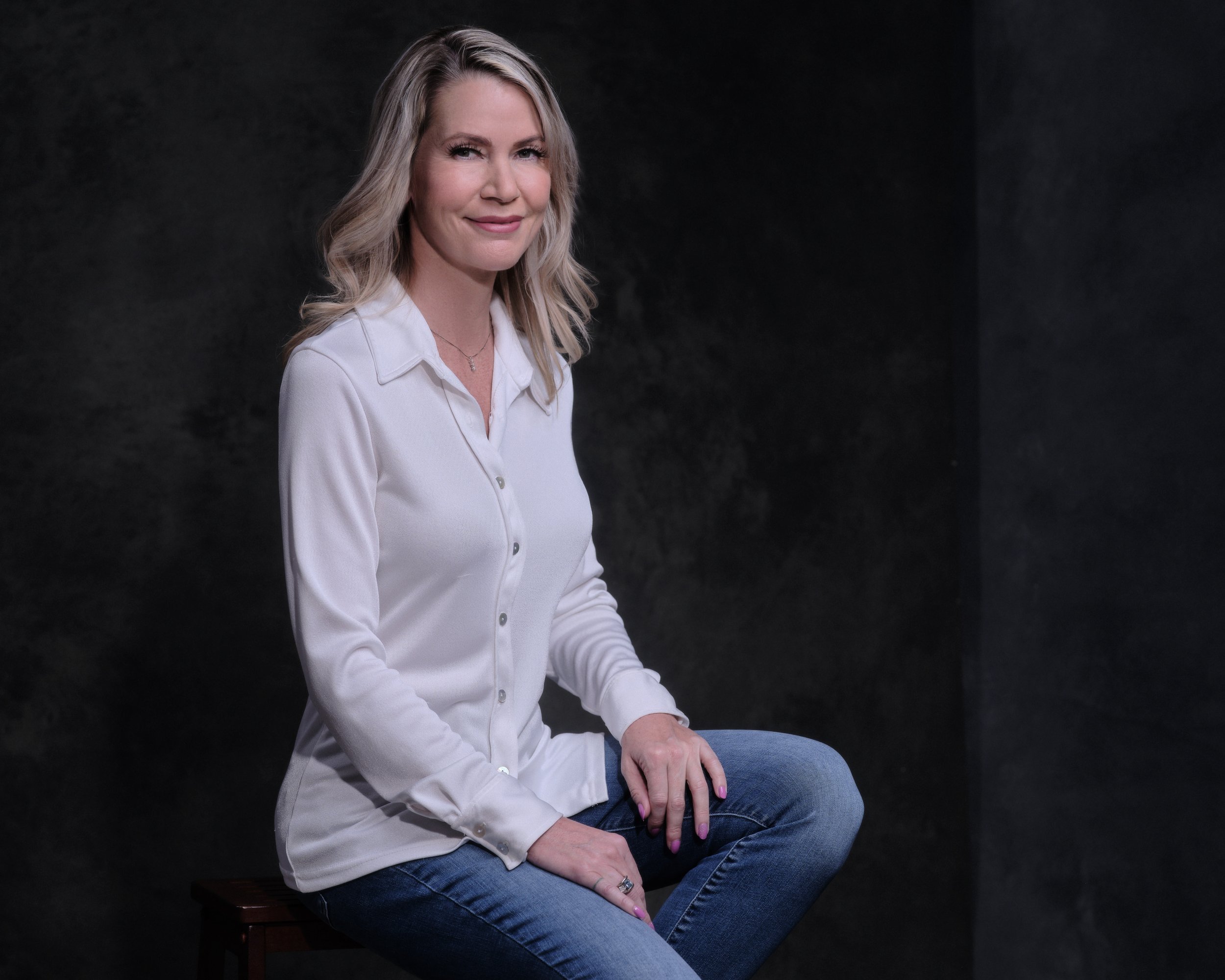 A woman with blonde wavy hair wearing a white blouse and blue jeans, sitting on a wooden stool against a dark background, looking at the camera.