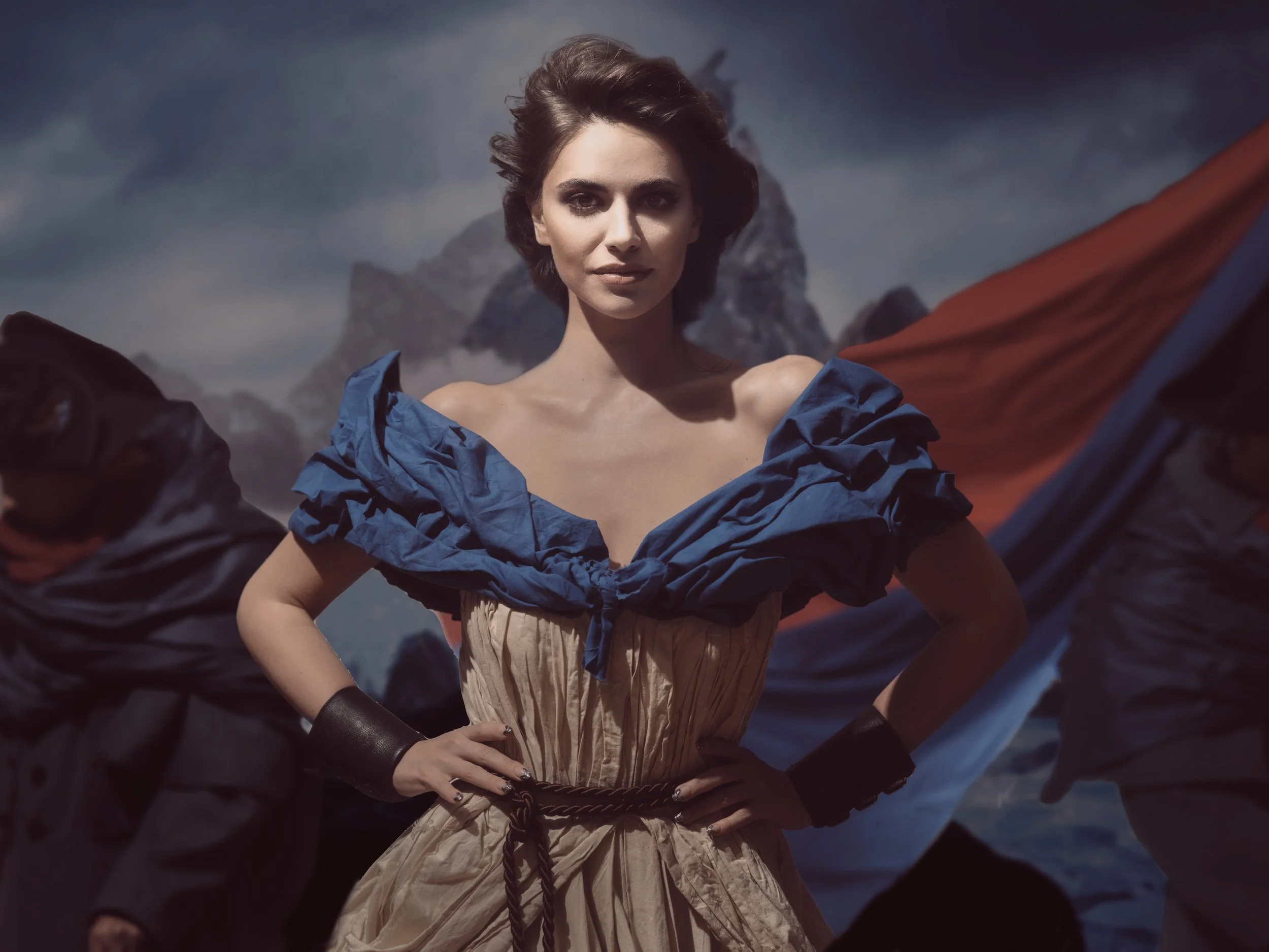 A woman wearing a ruffled blue and beige dress with a black belt stands confidently with her hands on her hips, flanked by two figures dressed in dark clothing and masks, against a mountainous background.