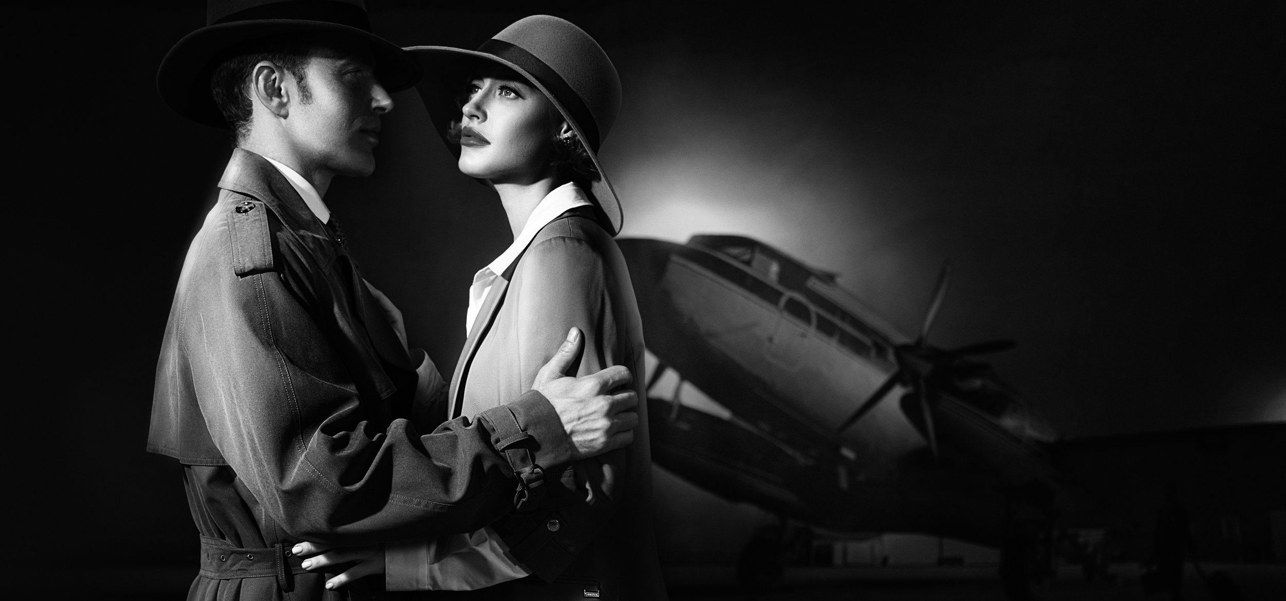 Black and white photograph of a man and woman in vintage military-style clothing, wearing wide-brimmed hats, standing close with the man's hand on the woman's shoulder, in front of a fighter jet.