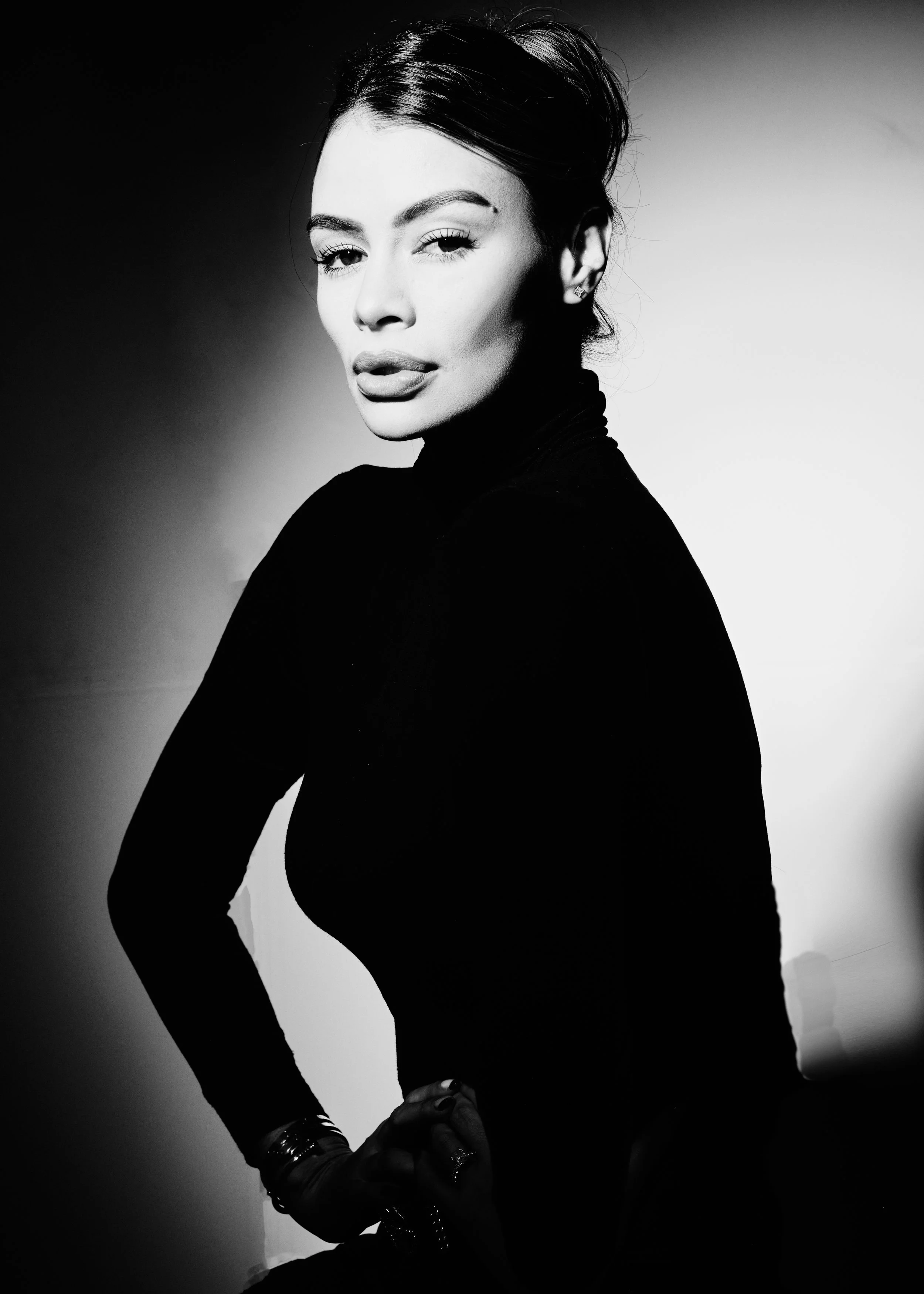Black and white portrait of a woman with stylized hair, wearing a high-neck top, lipstick, and jewelry, with one eye winking and a confident pose.