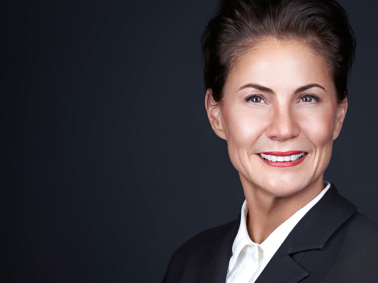 Professional portrait of a smiling woman with short dark hair, wearing a black blazer and white shirt, against a dark background.