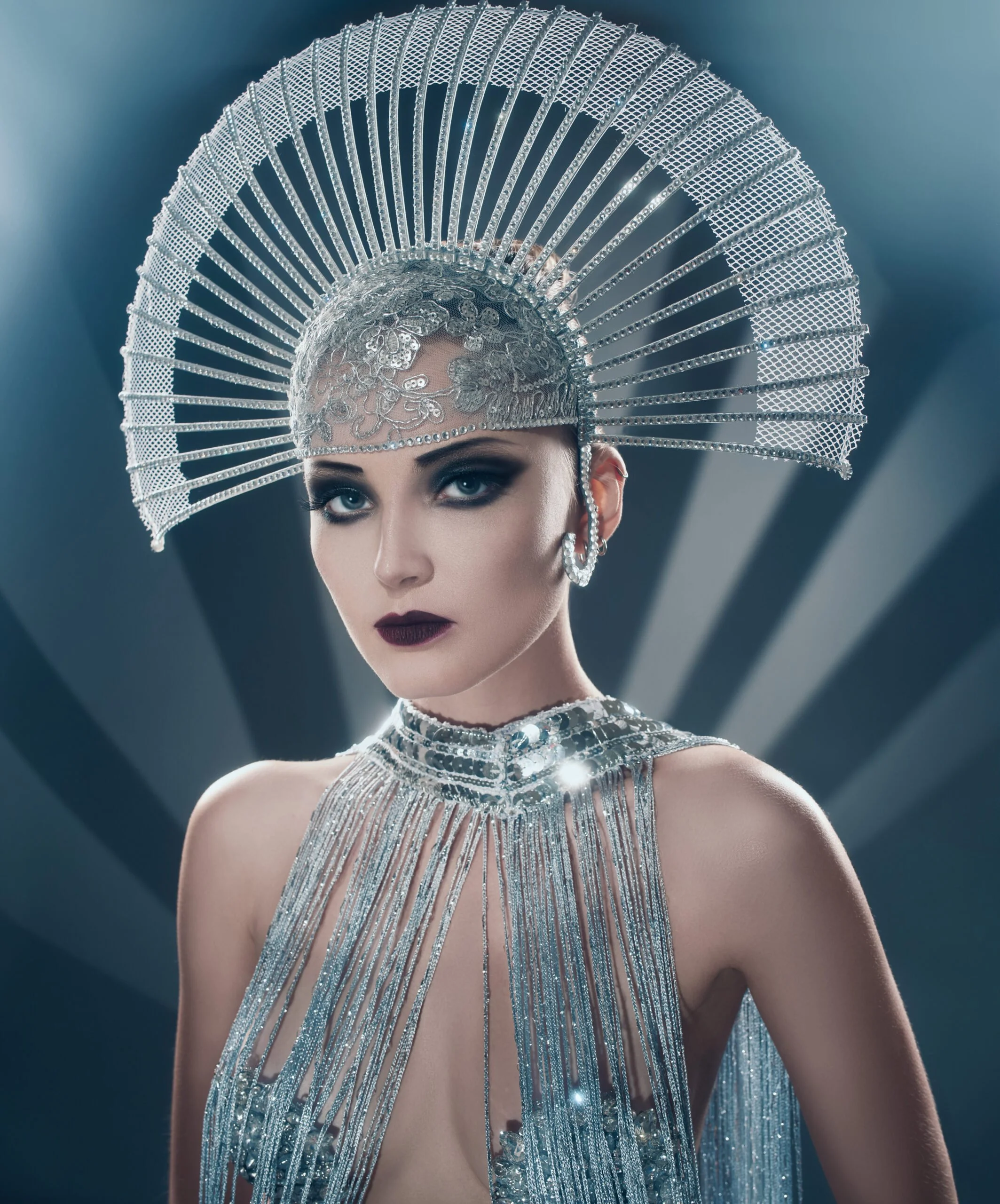 A woman wearing an elaborate silver headdress and jewelry, with dark makeup and a serious expression, against a stylized background.