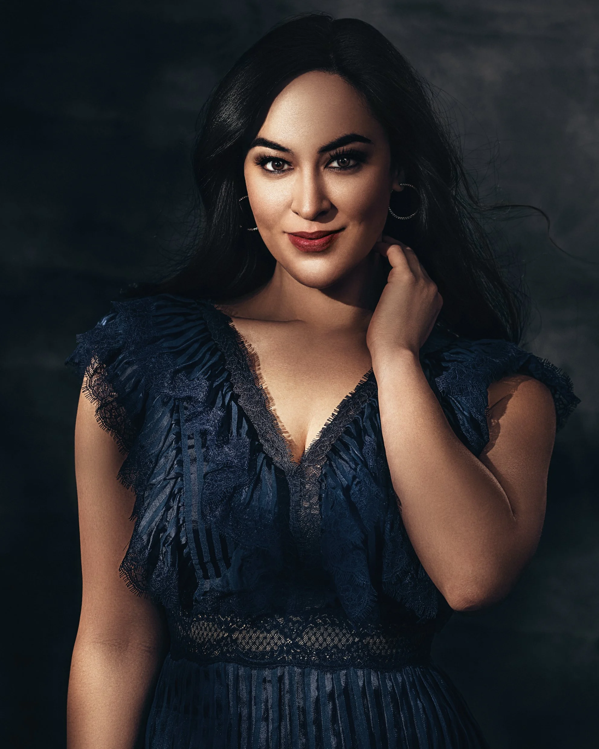 A woman with long dark hair wearing a dark blue lace dress, posing against a dark background.