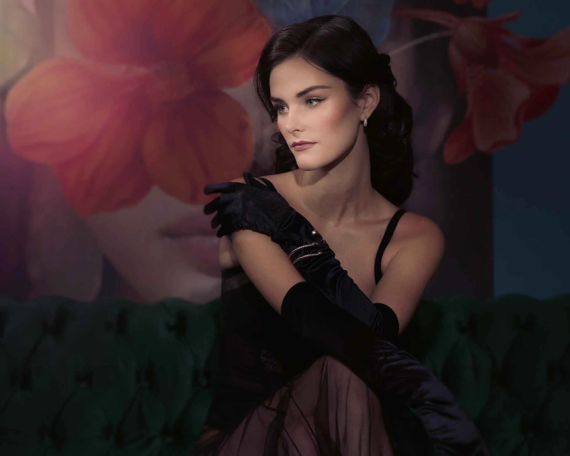 A woman with dark, wavy hair and fair skin, wearing a black dress with spaghetti straps and long black gloves, sitting on a dark green, tufted velvet sofa. She has a contemplative expression and is posing with her right hand on her left shoulder. In 