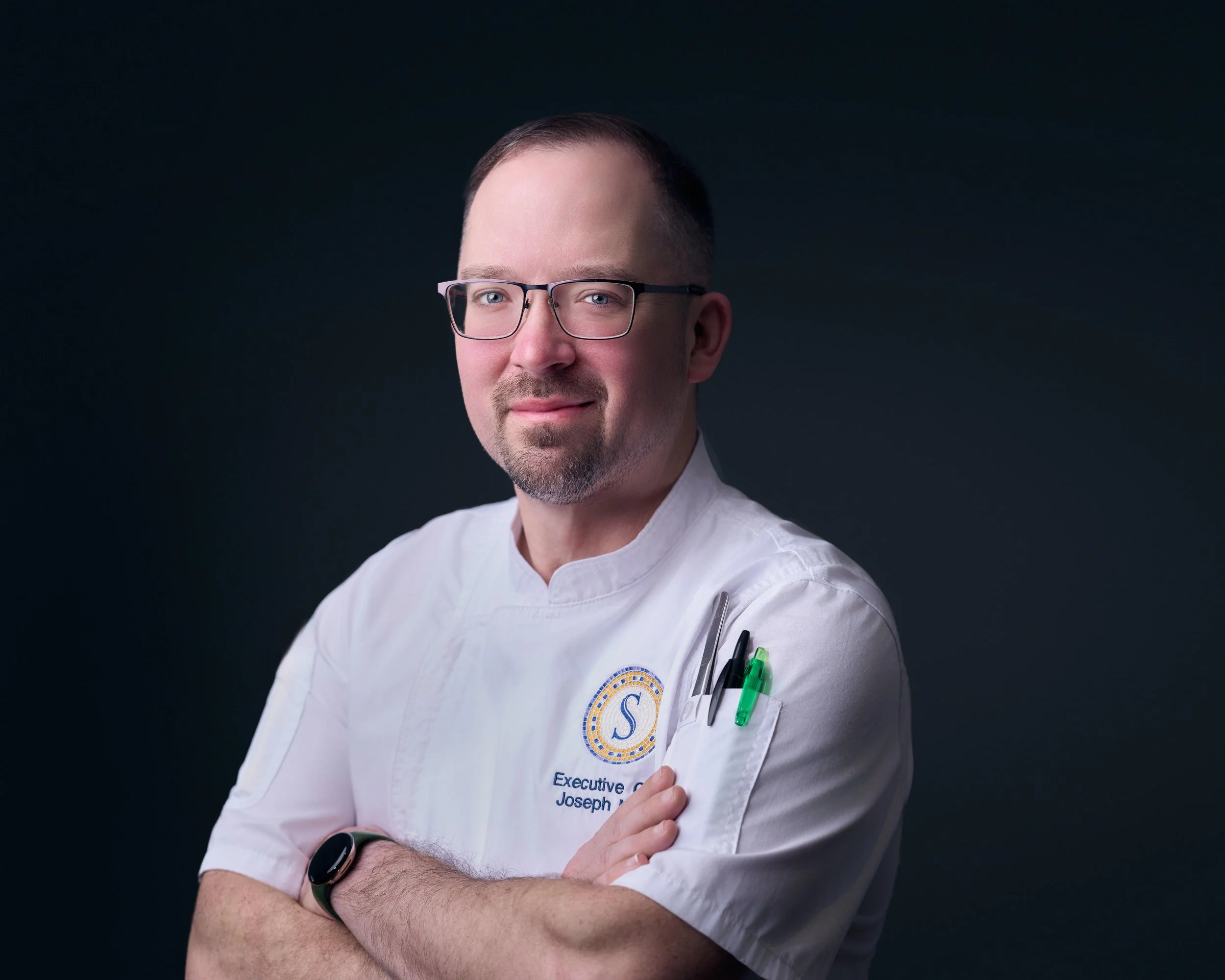 Portrait of a male chef wearing glasses and a white chef's coat, standing with arms crossed against a dark background.