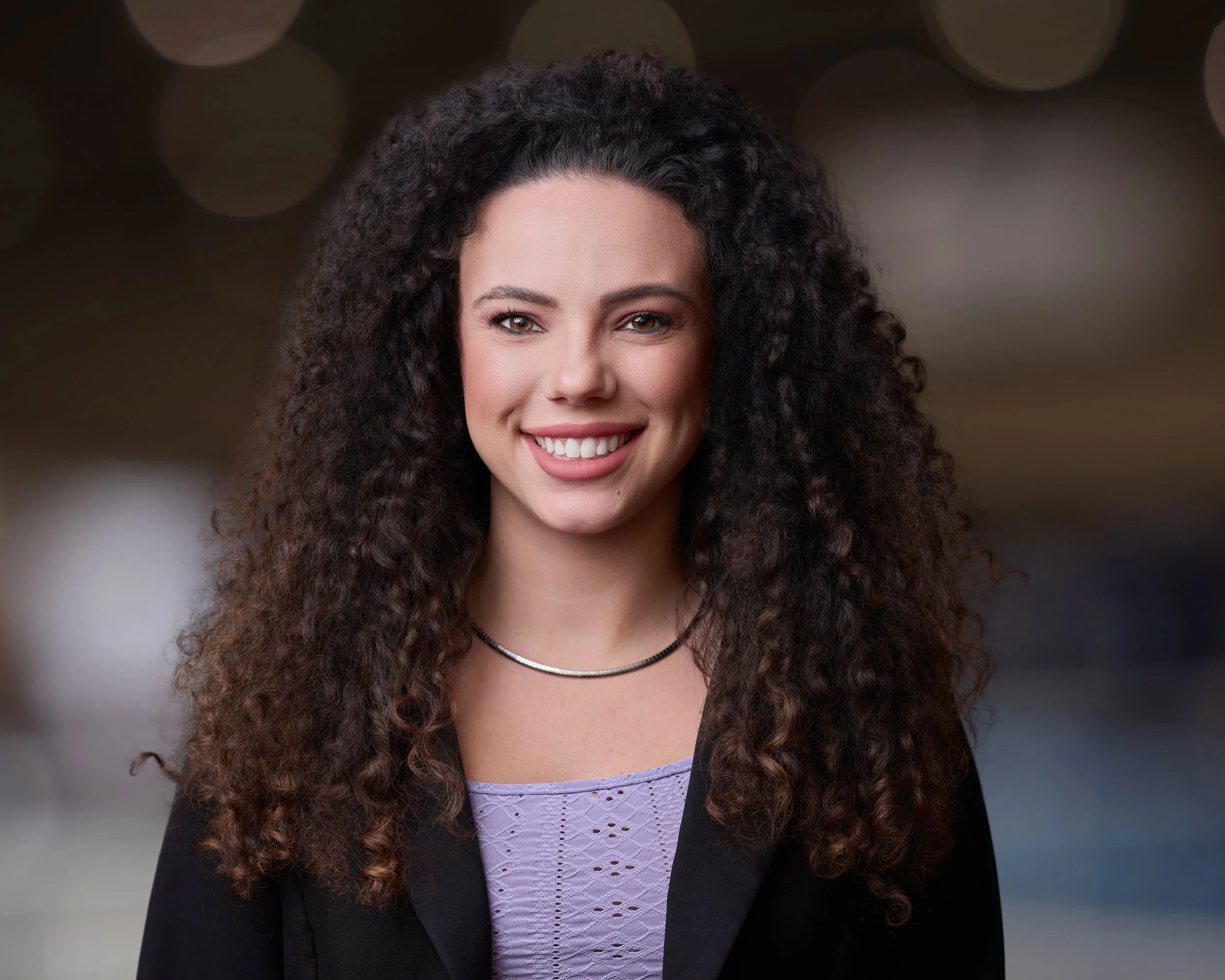 A young woman with long, curly dark hair smiling in a professional setting, wearing a black blazer and a light purple top with a necklace.
