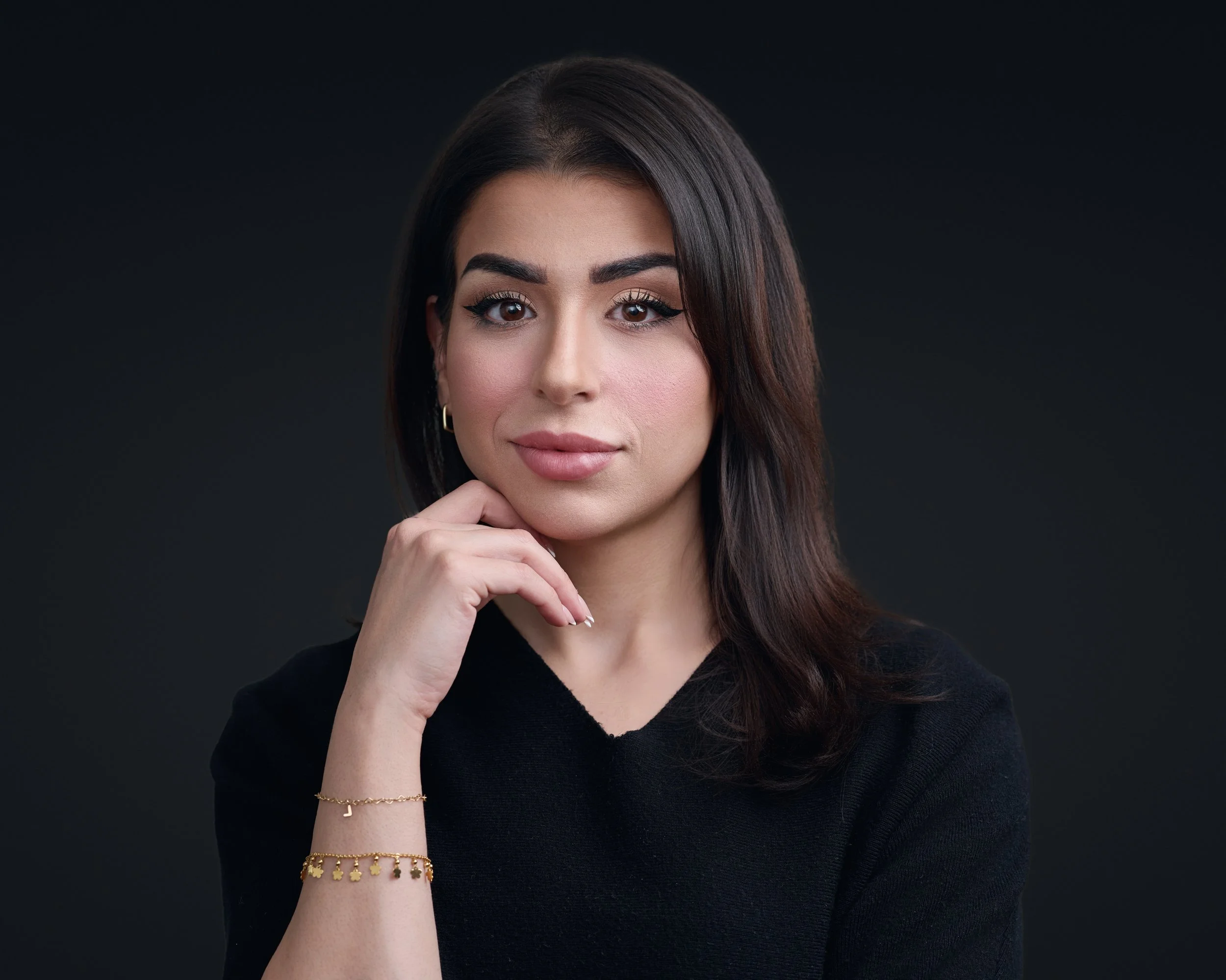 Portrait of a woman with dark hair and makeup, wearing a black top and gold jewelry, posing against a dark background.