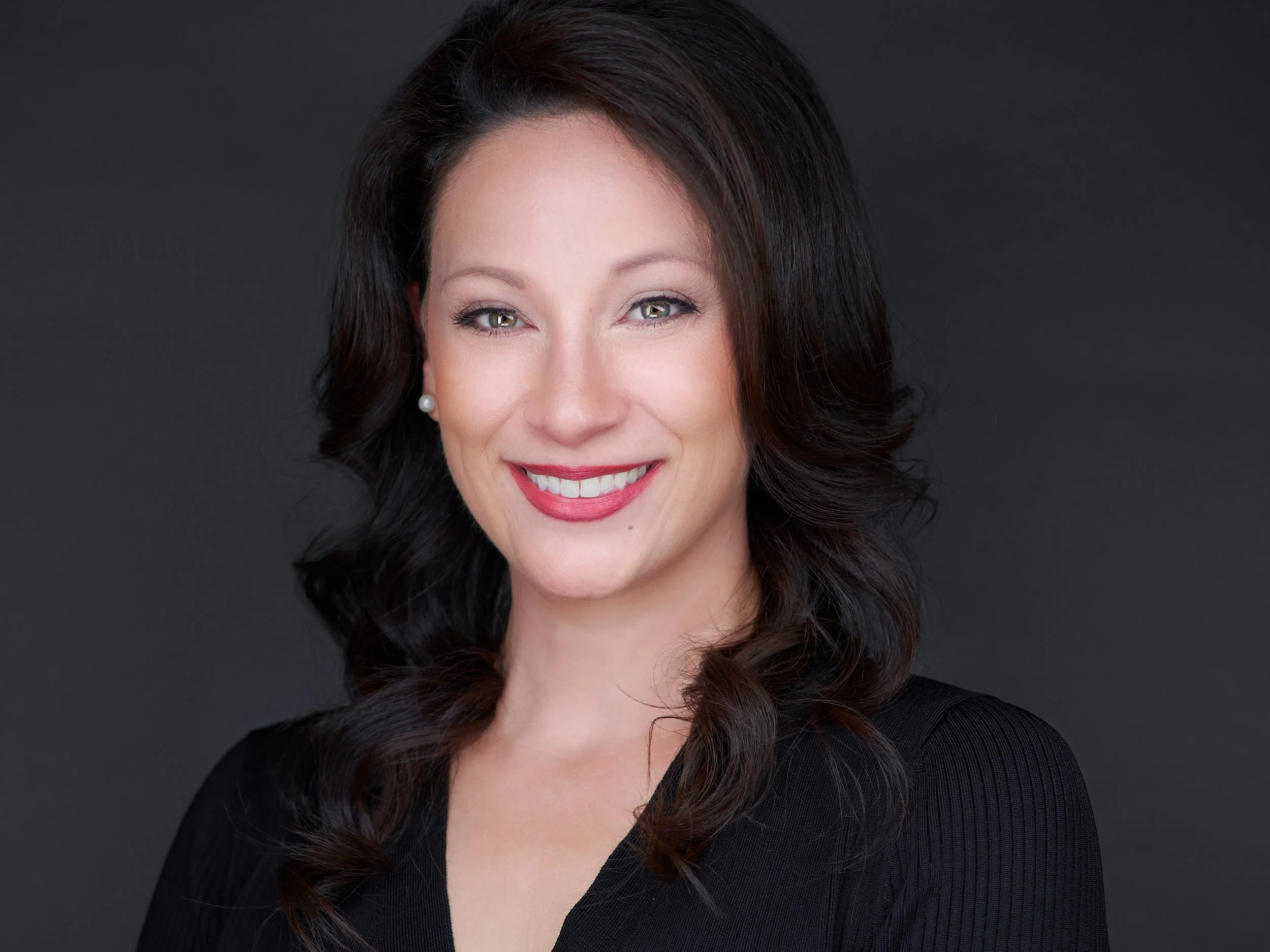 A woman with black wavy hair, wearing pearl earrings and a black top, smiling against a dark grey background.