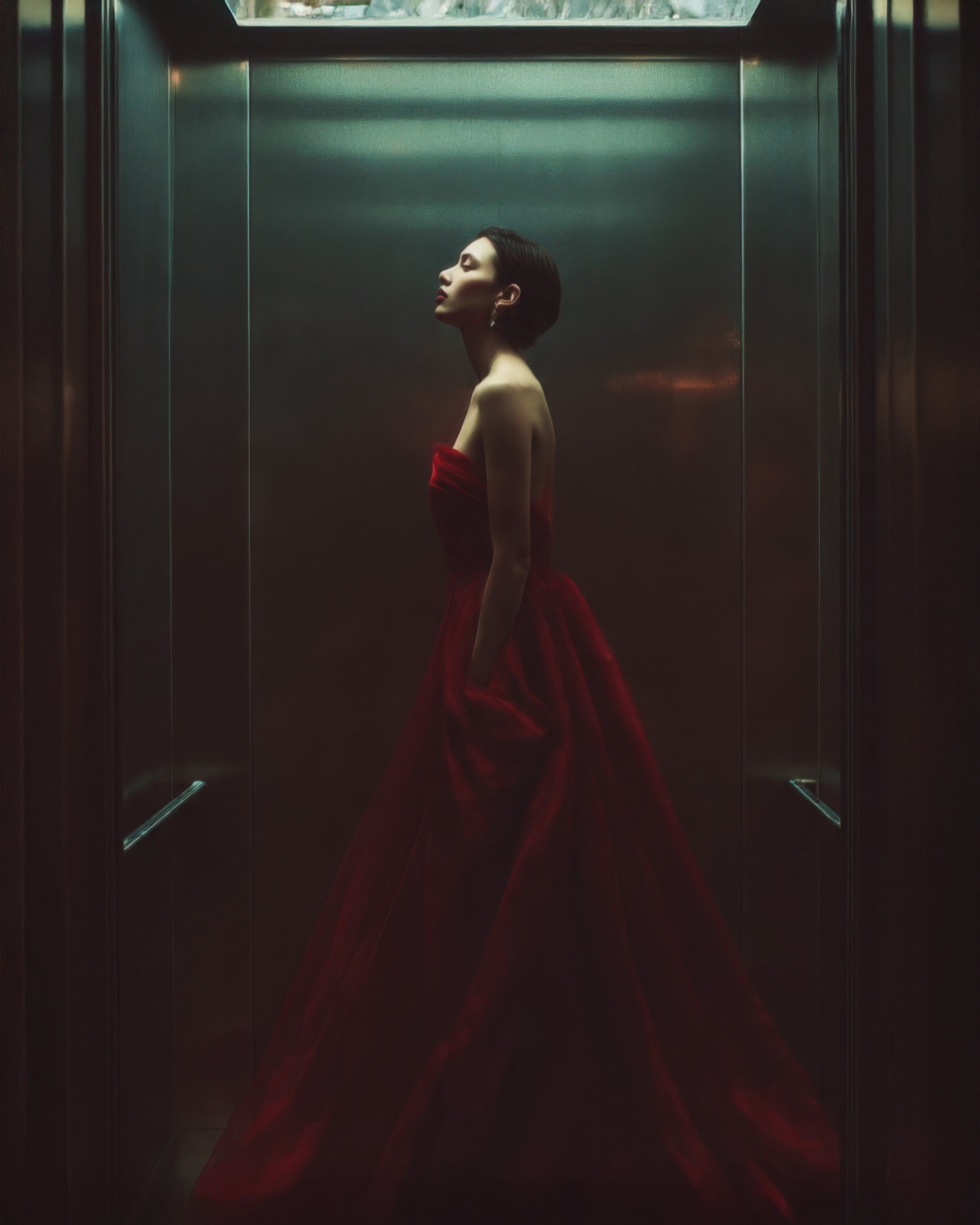 A woman in a red dress standing inside an elevator with metallic walls, facing left with her eyes closed.