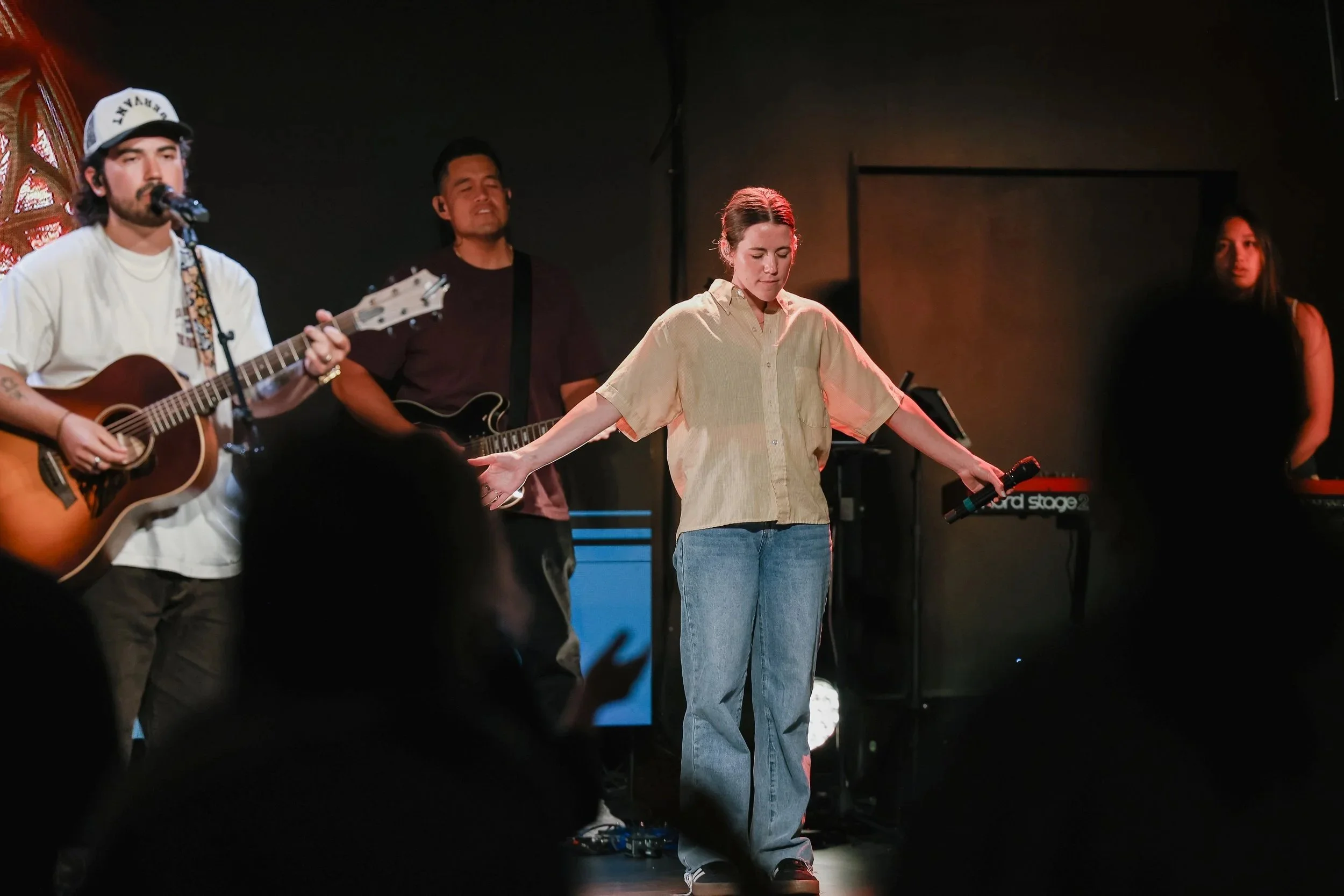 A band performs on stage with a stained glass window background, while the audience in the foreground raises their hands.