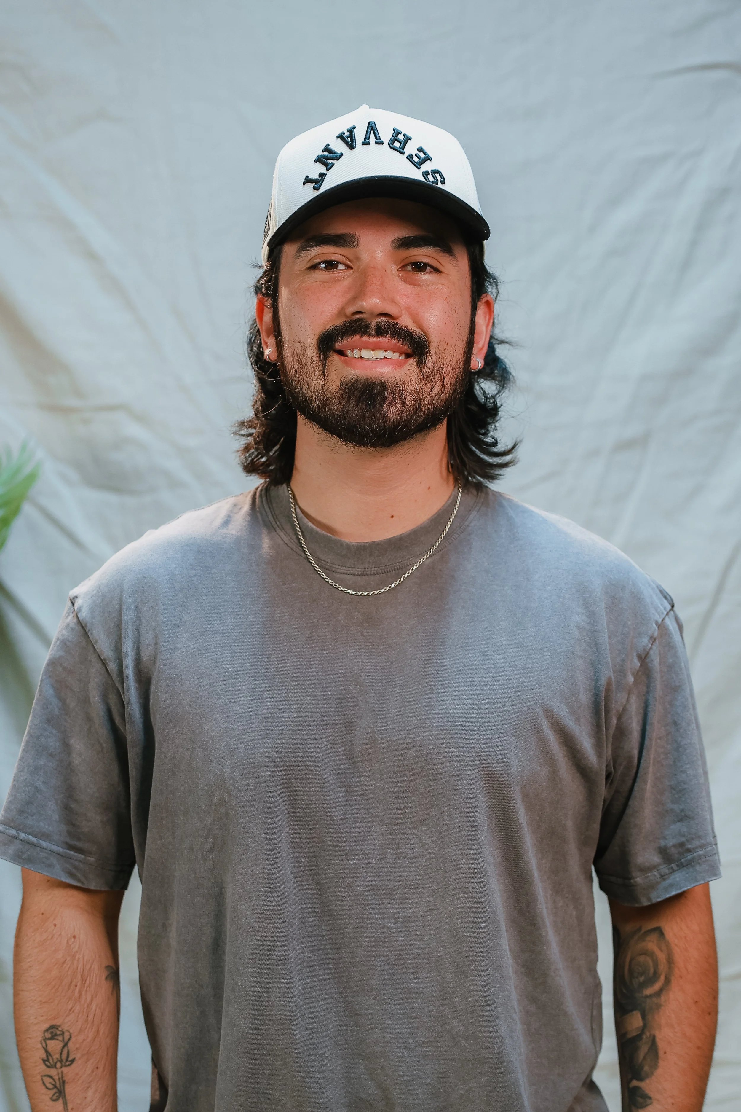 A smiling young man with a beard and tattoos, wearing a brown t-shirt, a chain necklace, and a baseball cap that says 'Plant seeds. Grow gardens.' standing against a plain light background.