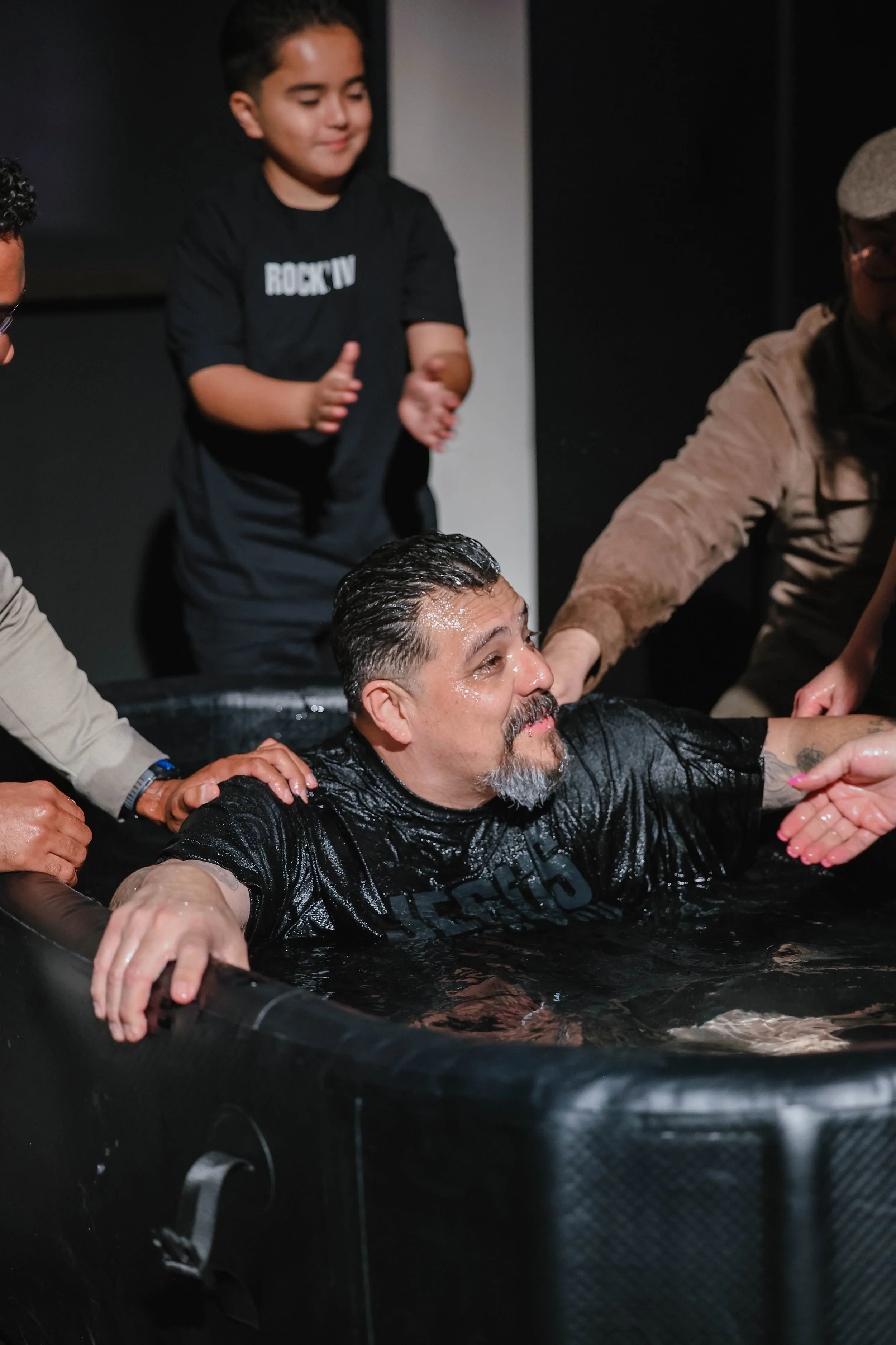 A man is submerged in water during a baptism ceremony, with his arms resting on the sides of a baptismal pool. He is surrounded by several people assisting him, and a young boy is standing behind him, clapping and smiling in support.