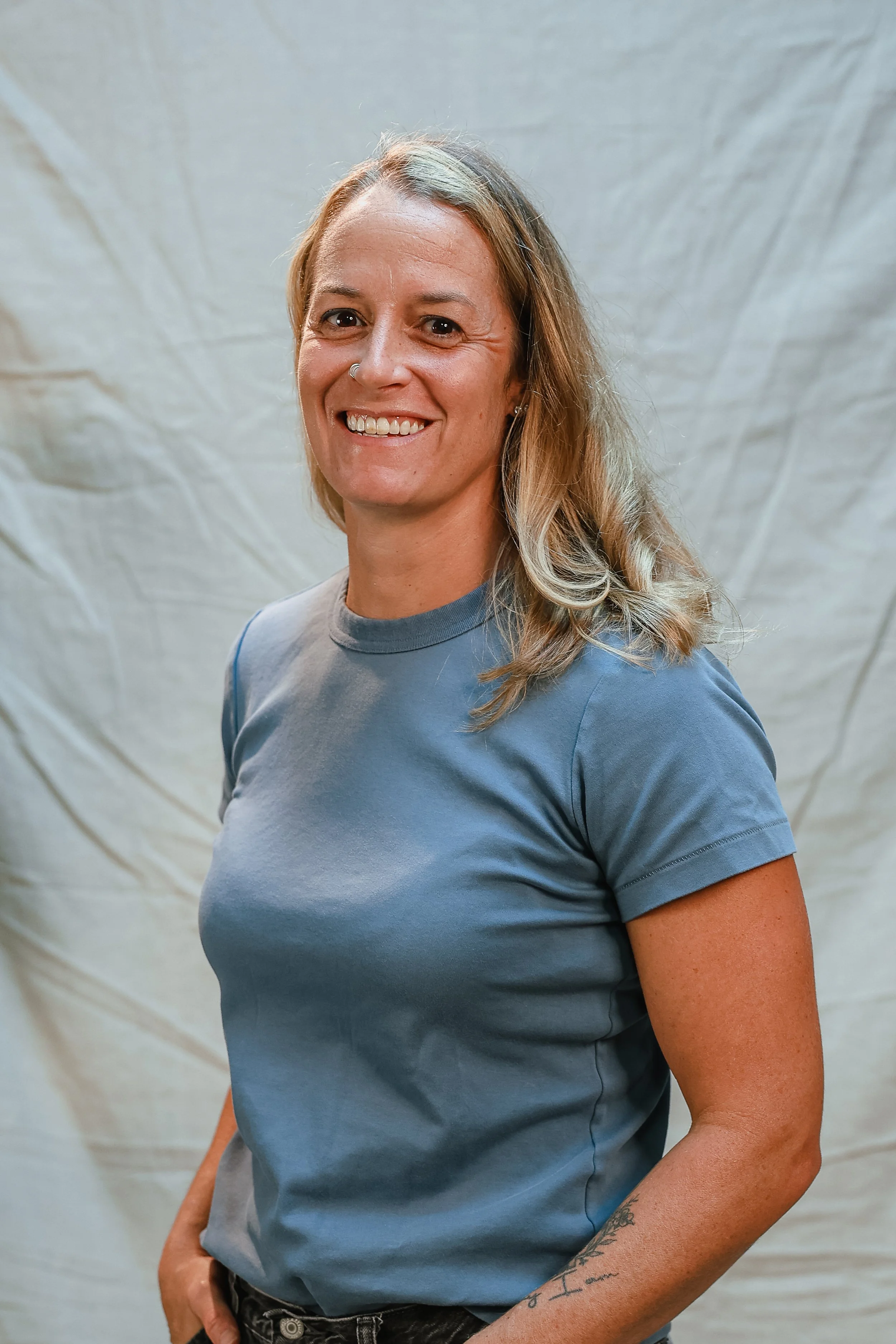 A woman with shoulder-length hair, smiling, wearing a white t-shirt, beige pants, and a smartwatch, standing with arms crossed against a plain light-colored background.