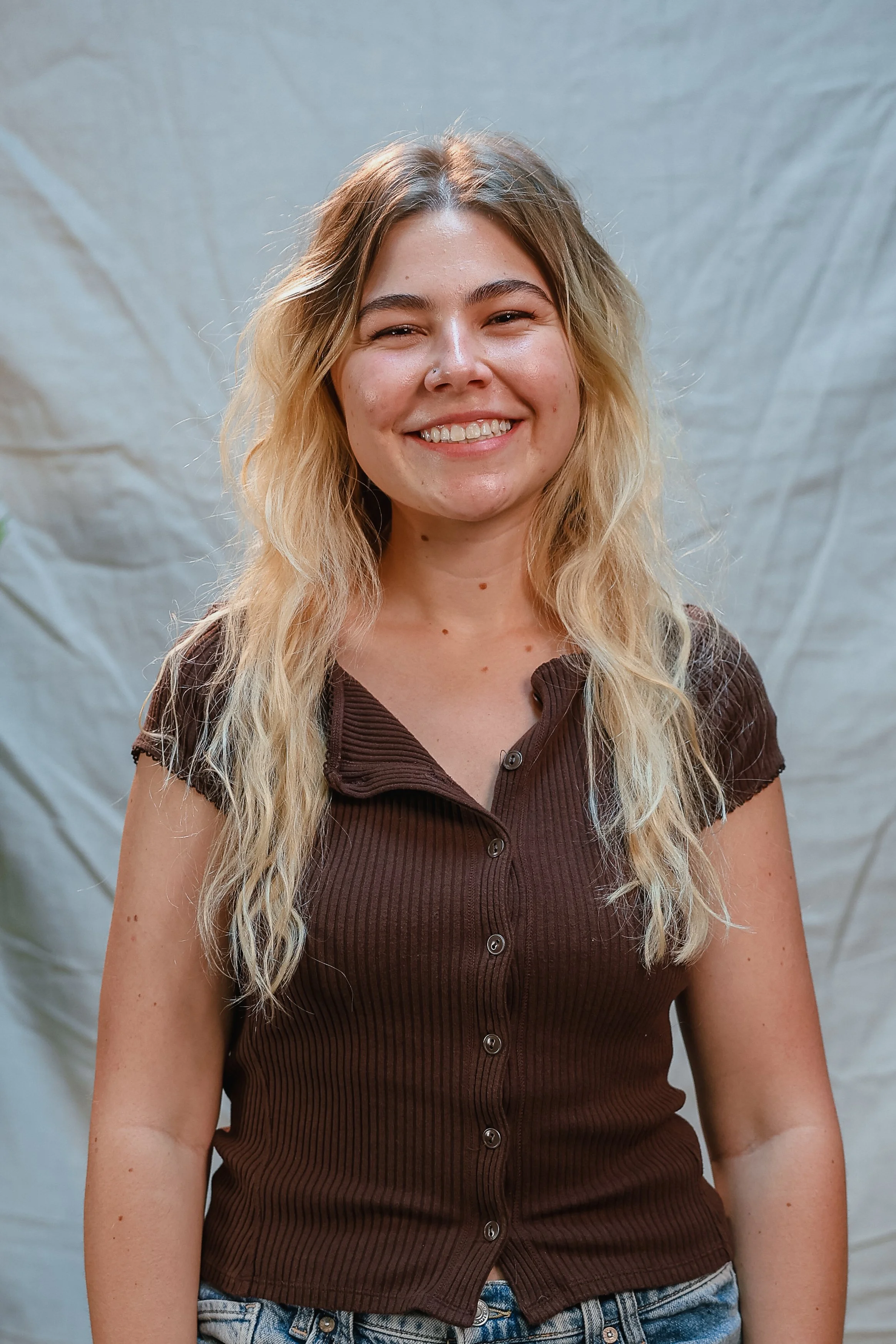 A smiling young woman with long, wavy blonde hair wearing a white t-shirt and white overalls, standing against a plain light-colored background.