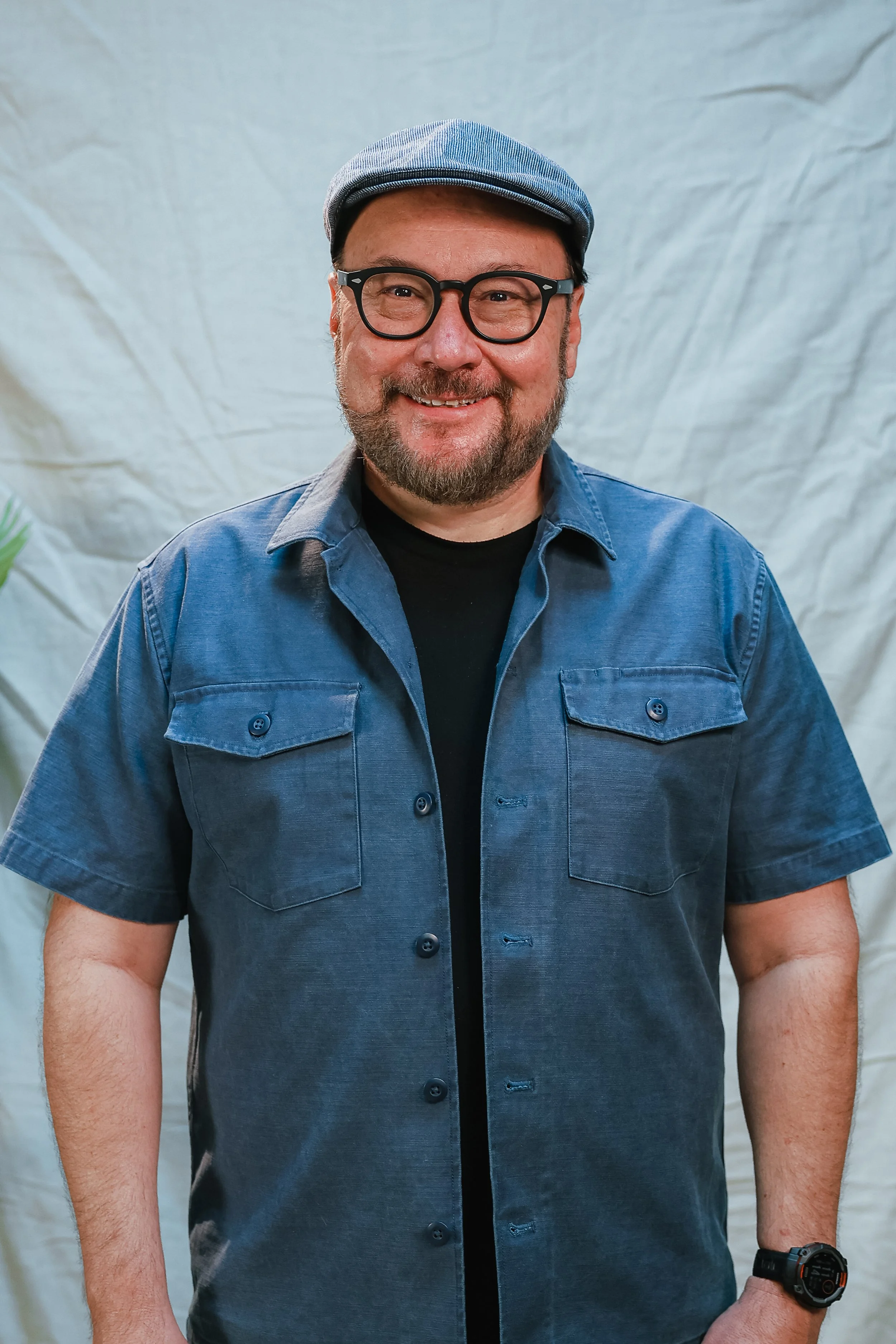 A smiling middle-aged man wearing glasses, a gray cap, a light gray short-sleeve button-up shirt over a dark t-shirt, standing against a plain white background.