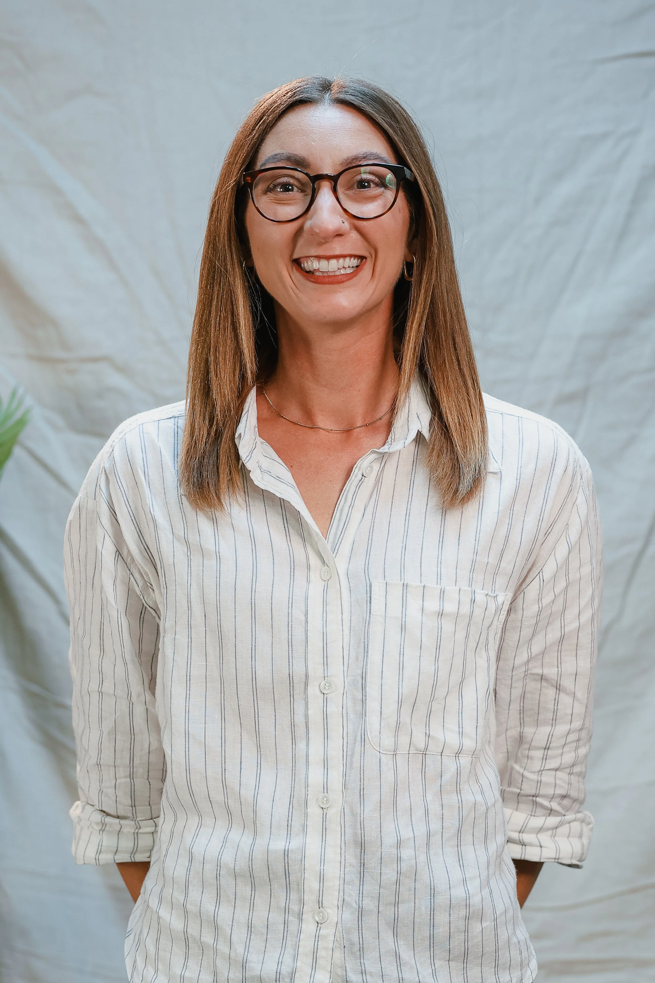 A woman with shoulder-length wavy brown hair, glasses, and a big smile, standing against a plain light-colored wall, wearing a brown jacket over a white t-shirt and blue jeans.
