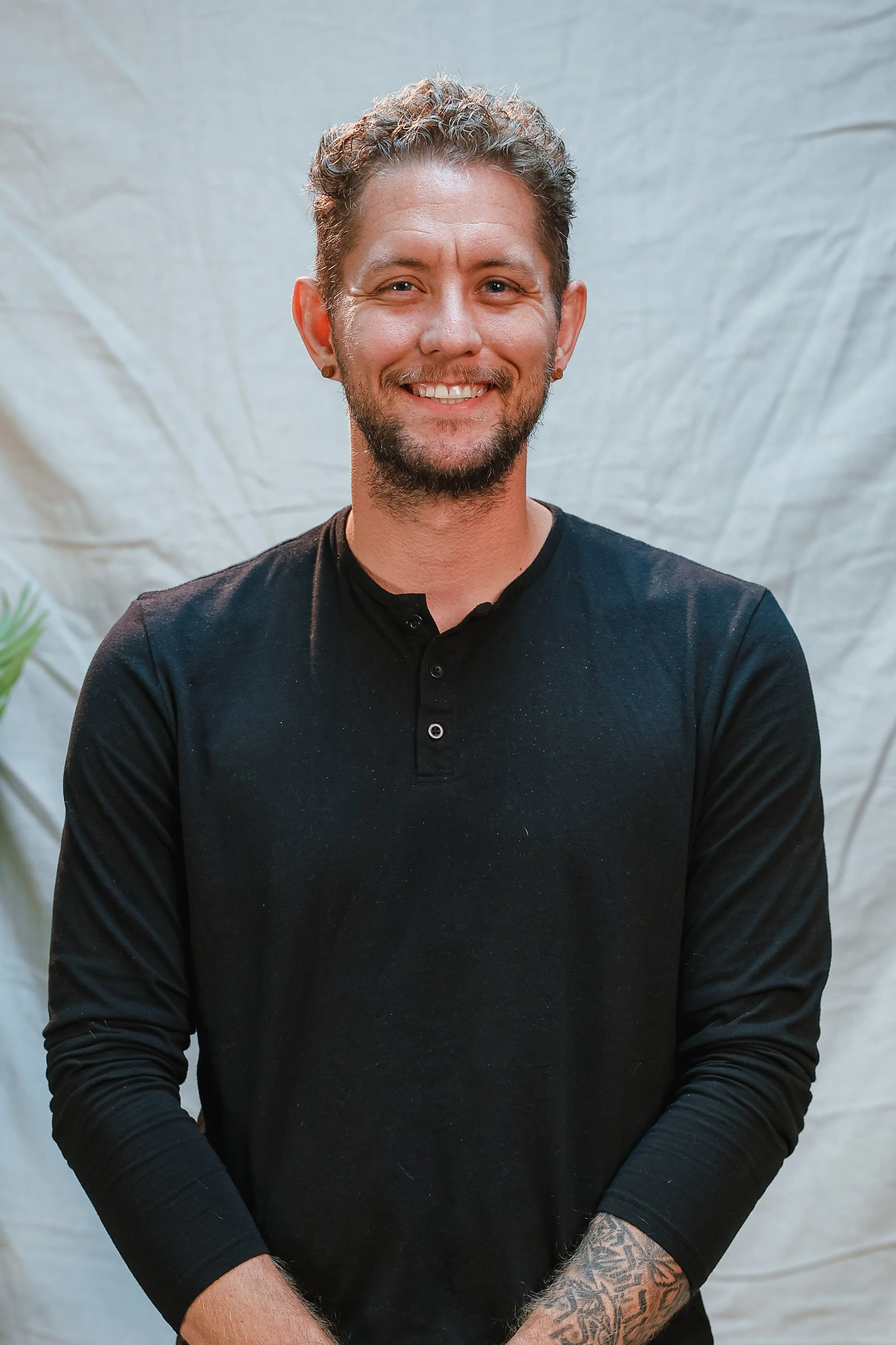Smiling man with curly hair and tattoos wearing a white shirt