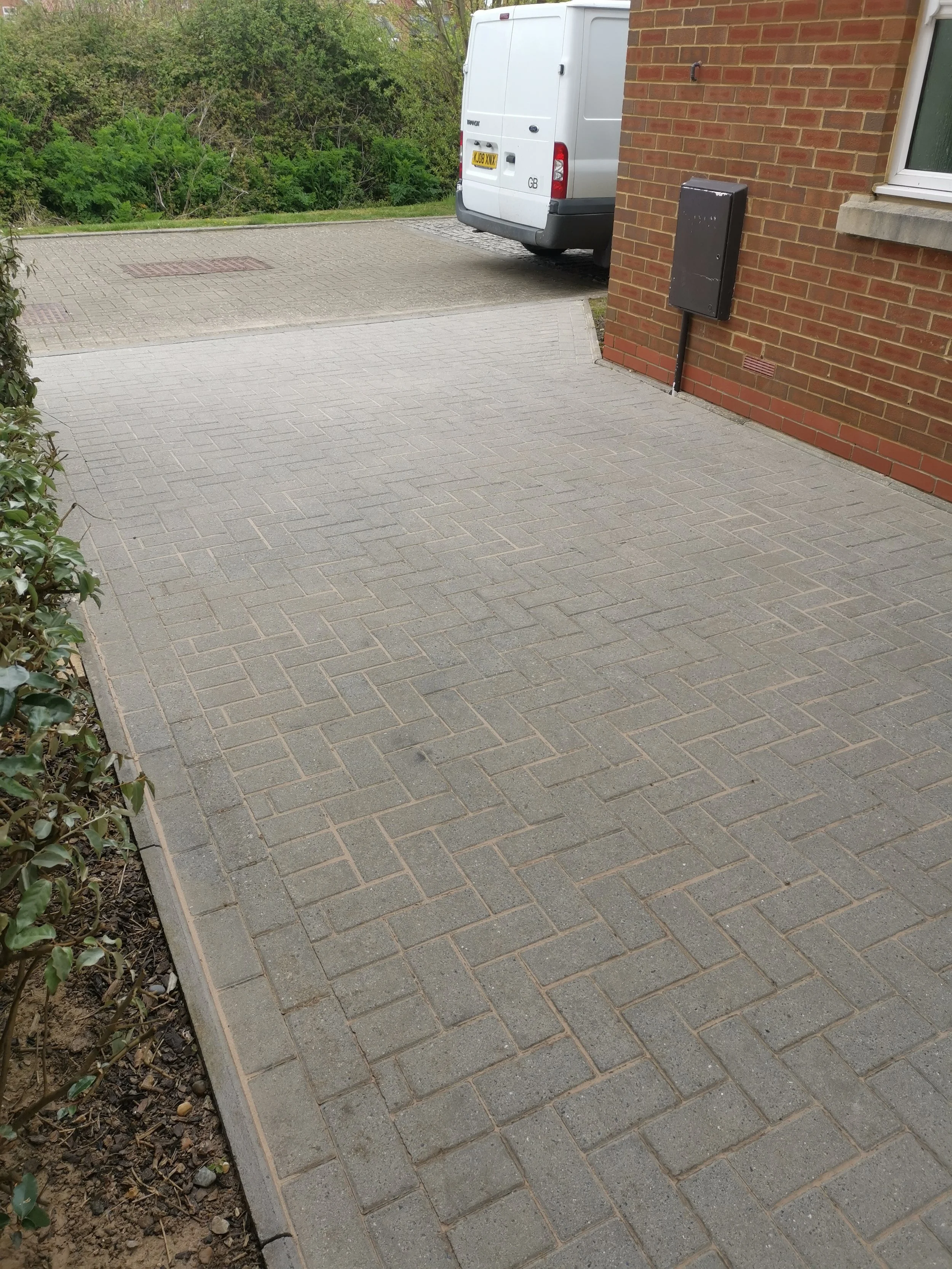 A paved walkway next to a brick house with a black utility box on the wall, a white van parked nearby, and a grassy area with trees in the background.