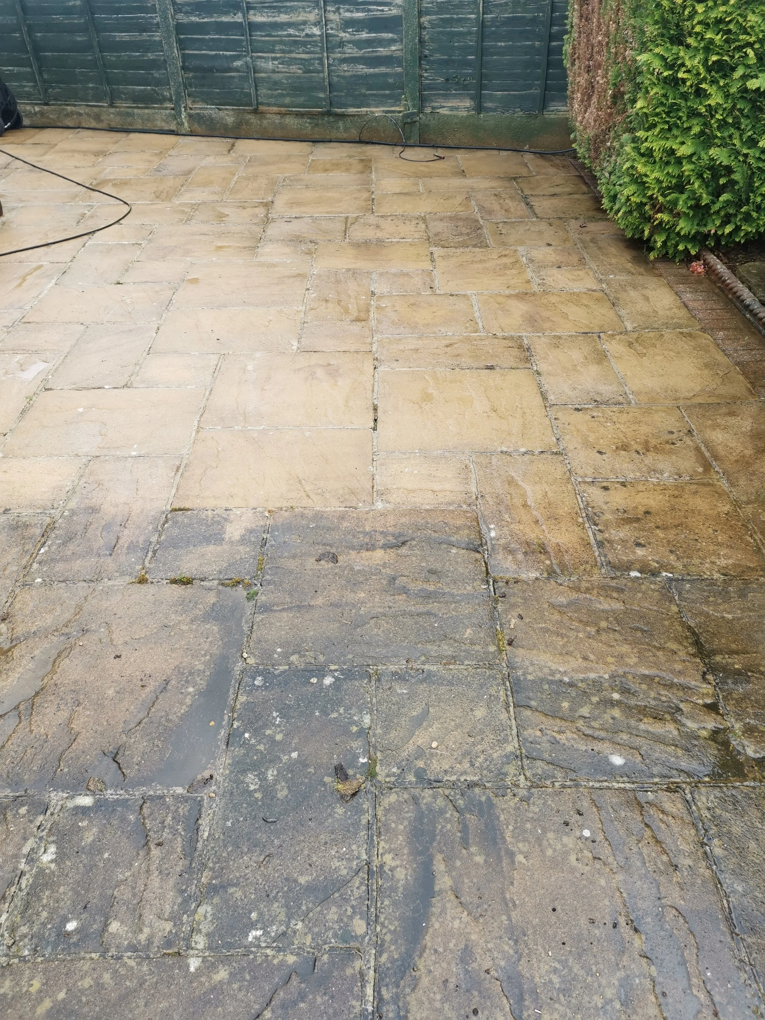 Clean and wet paved stone patio with a green wooden fence and bush in the background.