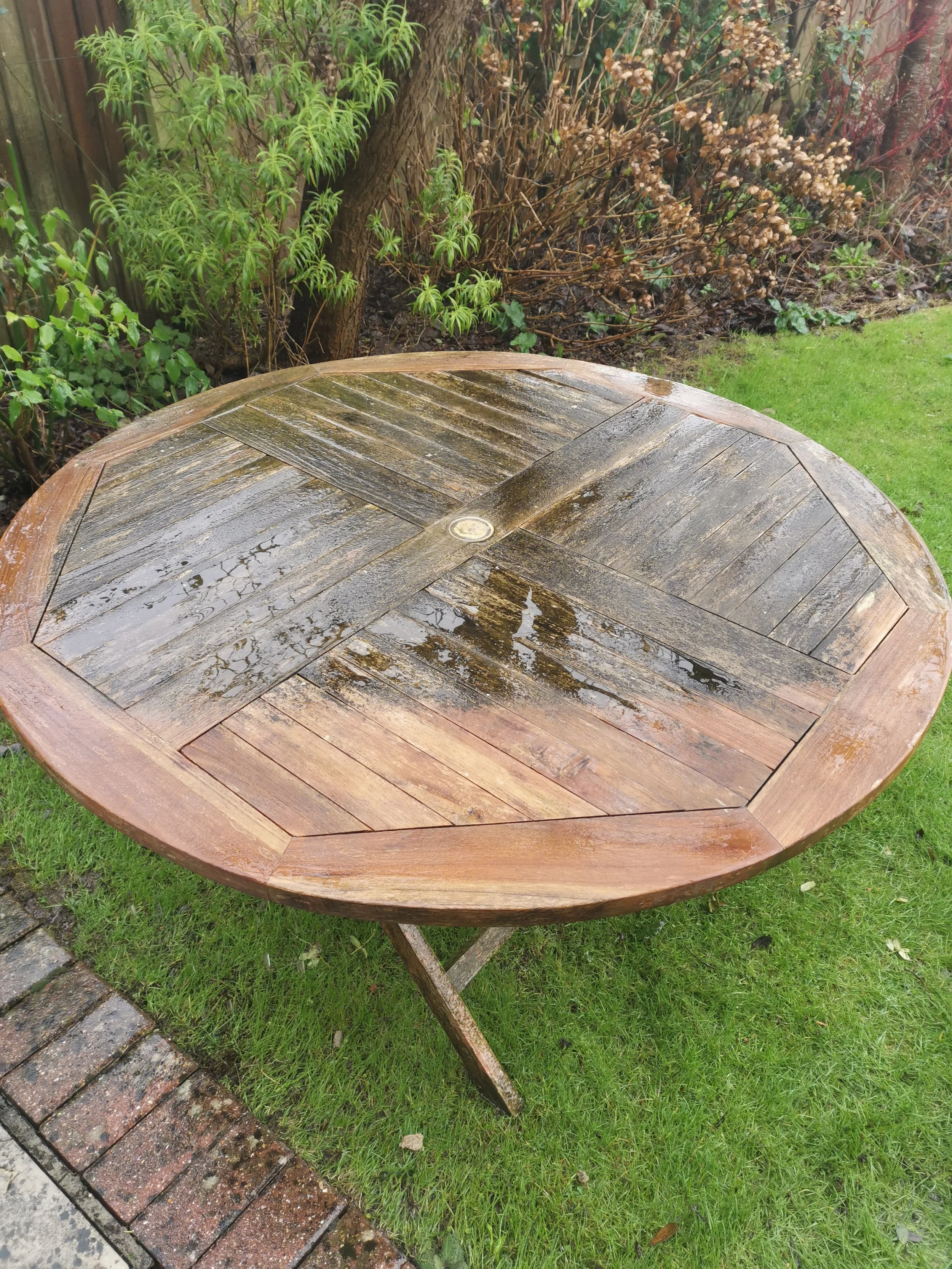 Wet wooden round outdoor table with a central metal bolt, situated on a grassy area next to a brick path and garden with shrubs and trees.