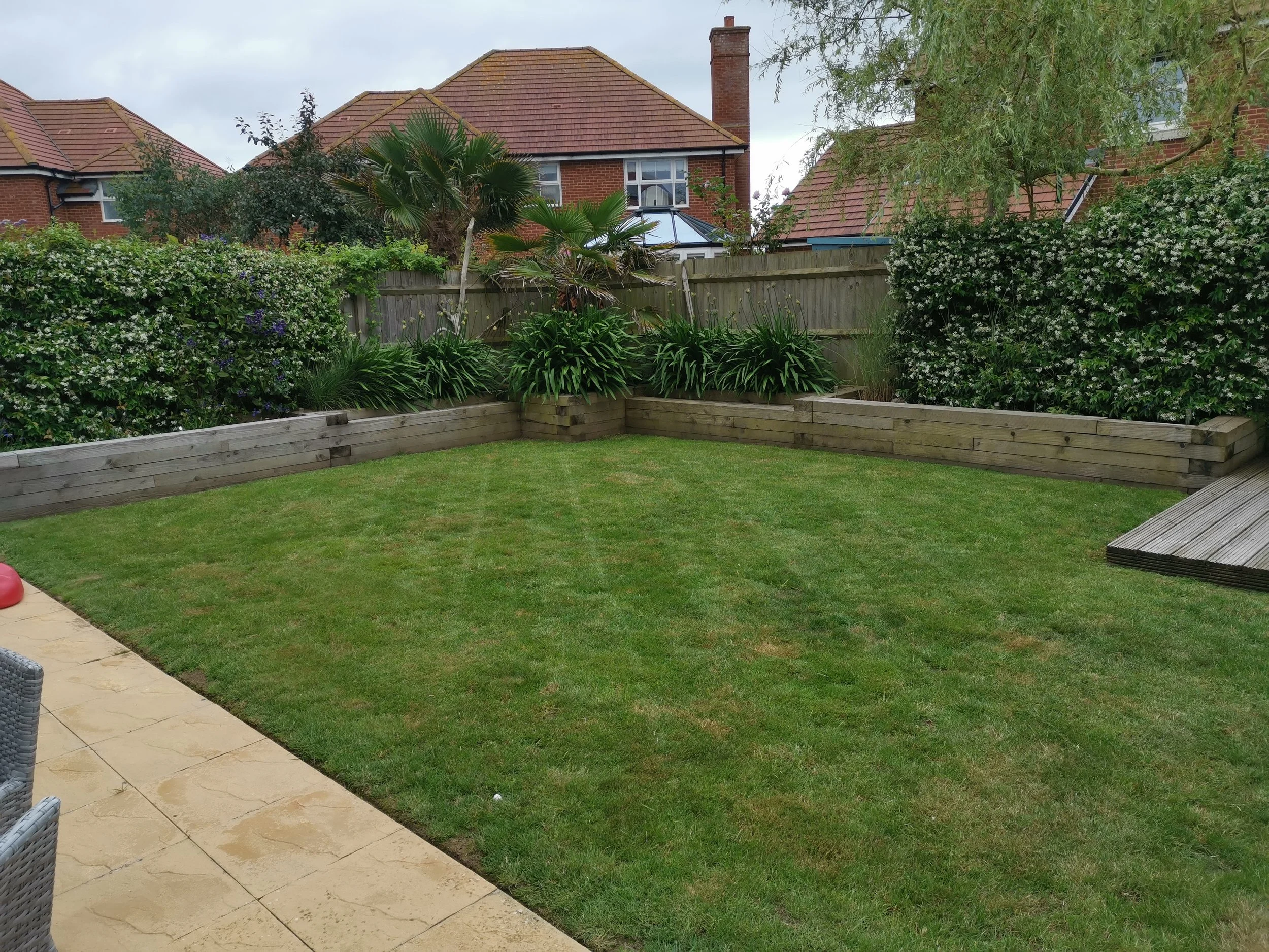 A backyard with well-maintained green grass, a stone patio with outdoor chairs, wooden garden beds with plants, and a tall wooden fence. There are several trees and flowering bushes in the background.