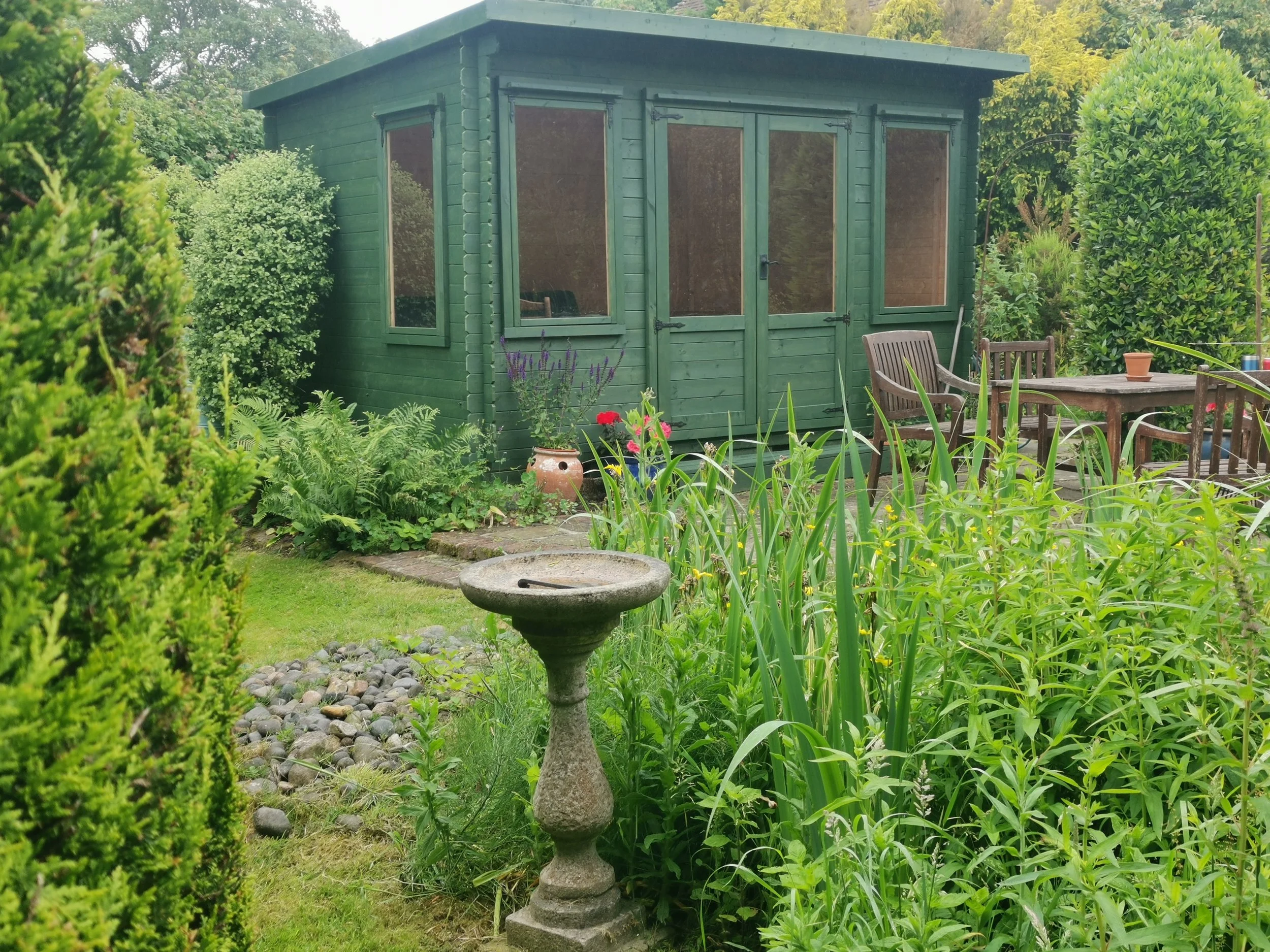 A green garden shed surrounded by lush plants and shrubs, with a stone birdbath and a wooden outdoor table with chairs.