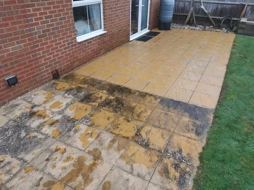 A backyard patio with partially cleaned and newly installed tiles next to a brick house wall, a window, and a grassy area.